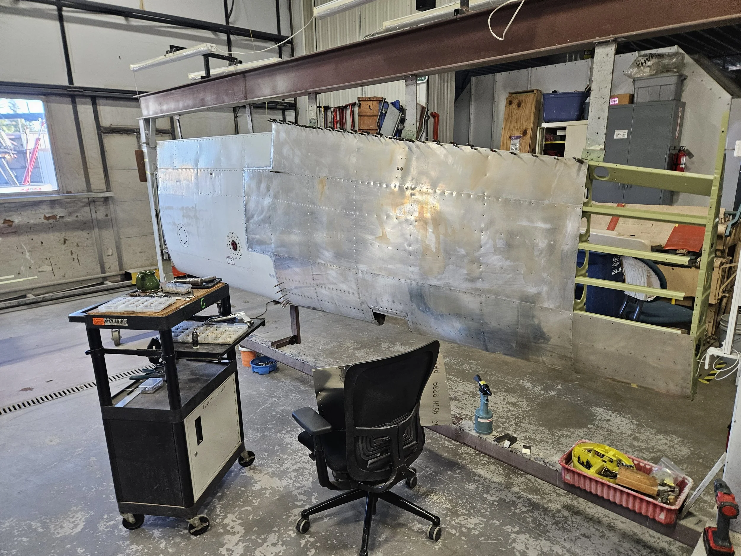 An indoor workshop with a partially assembled aircraft fuselage, surrounded by tools, workbenches, and storage cabinets.