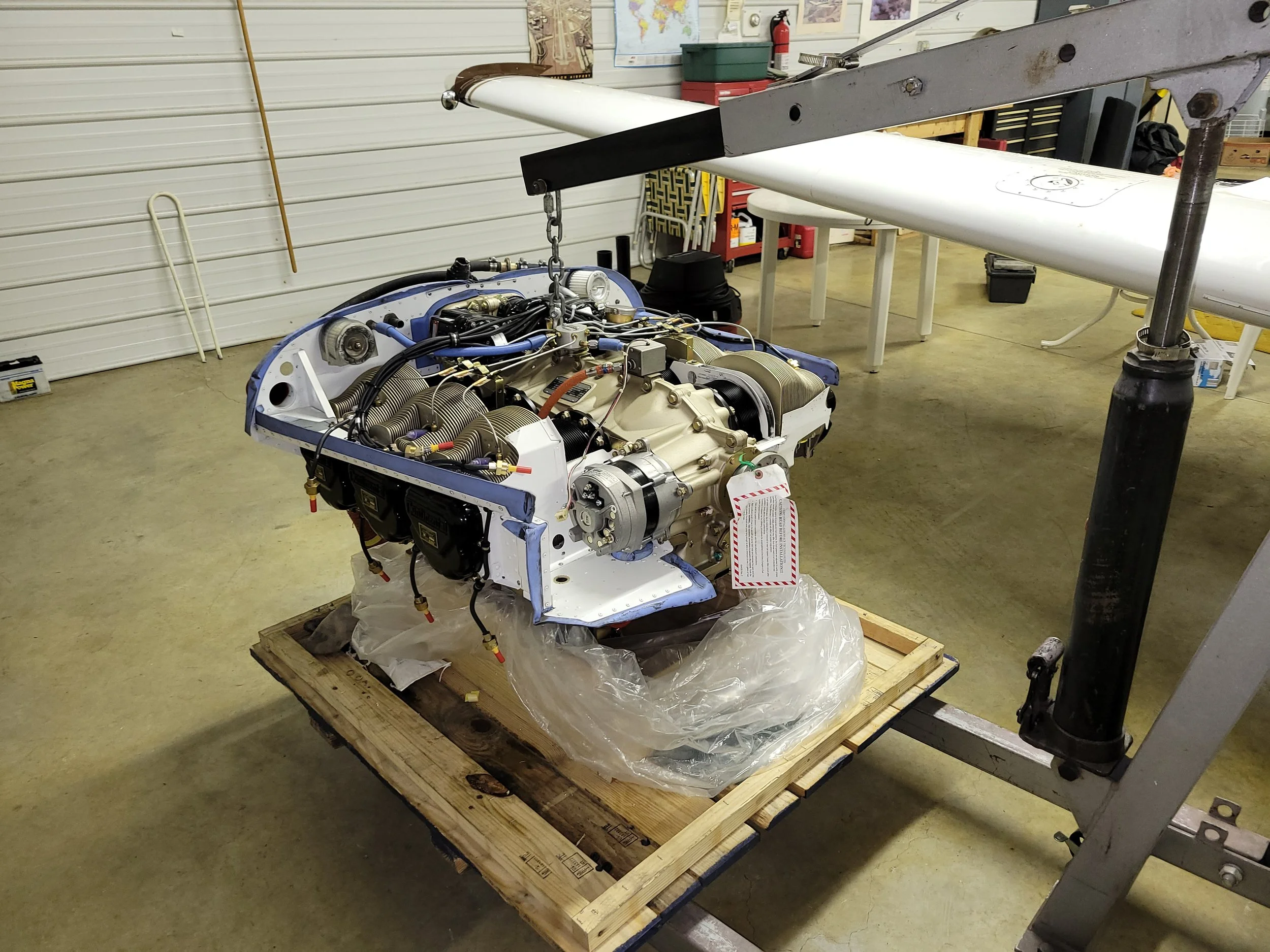 A partially assembled aircraft engine resting on a wooden pallet inside a workshop. Overhead, a wing is supported by a stand, and tools and storage are visible on the walls.
