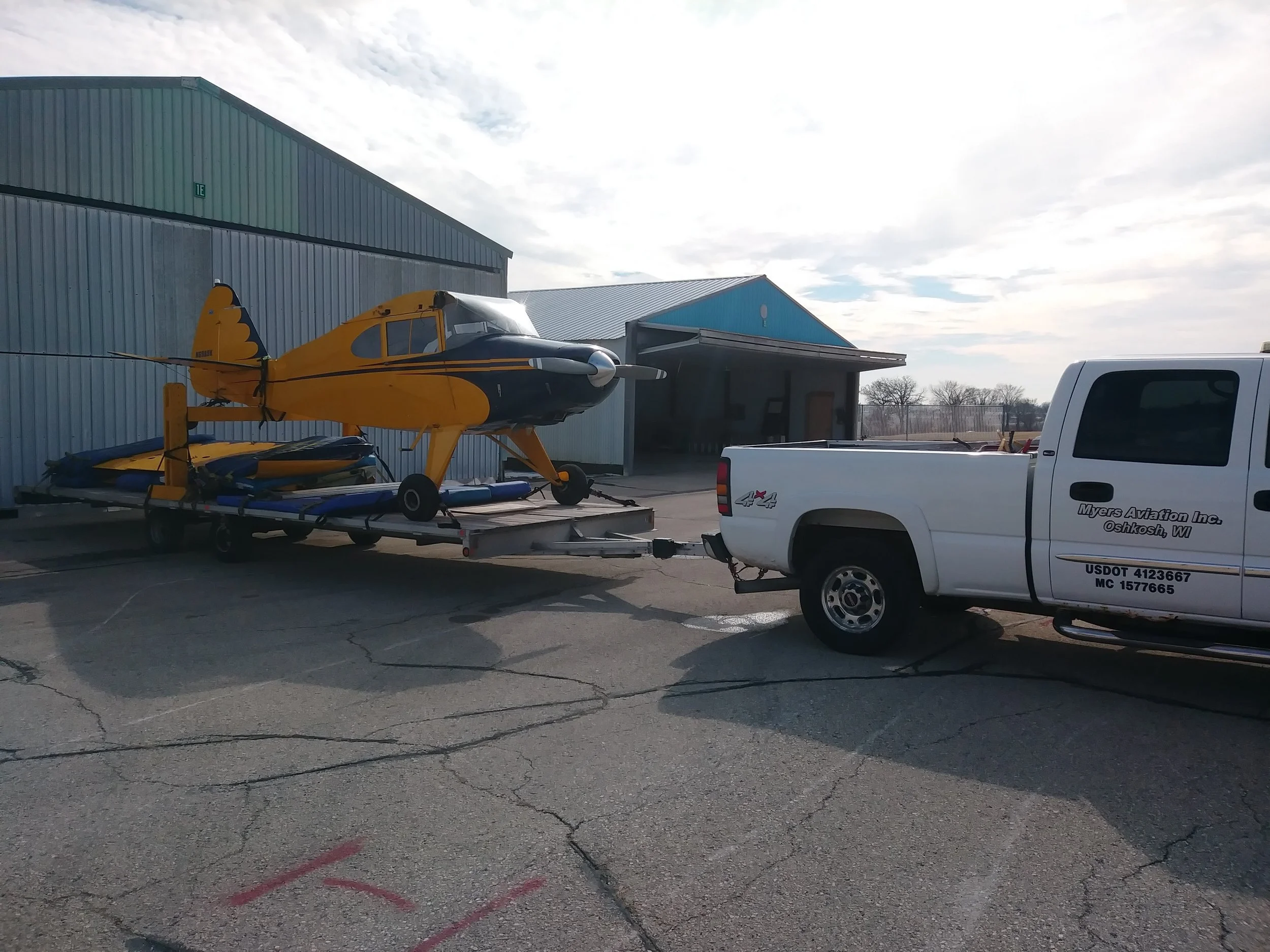 A yellow and black aircraft on a trailer, parked outside near a large metal building with a small structure attached, with a pickup truck nearby, under a partly cloudy sky.