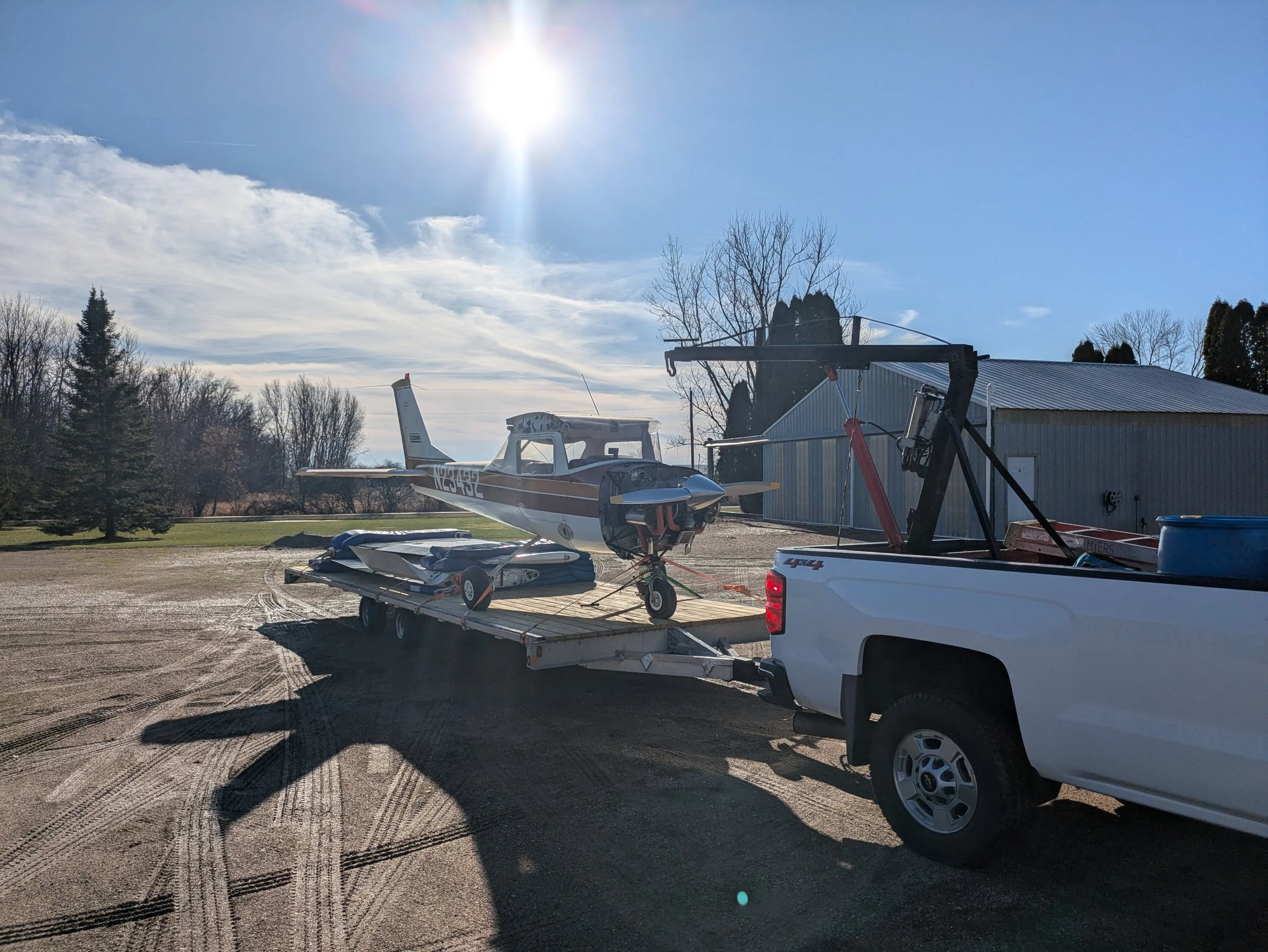 A small propeller airplane is on a trailer attached to a white pickup truck. The airplane appears to be in the process of being transported or stored at an outdoor location with a gravel surface. The background includes a blue sky with some clouds, a metal shed or hangar, and some leafless trees.