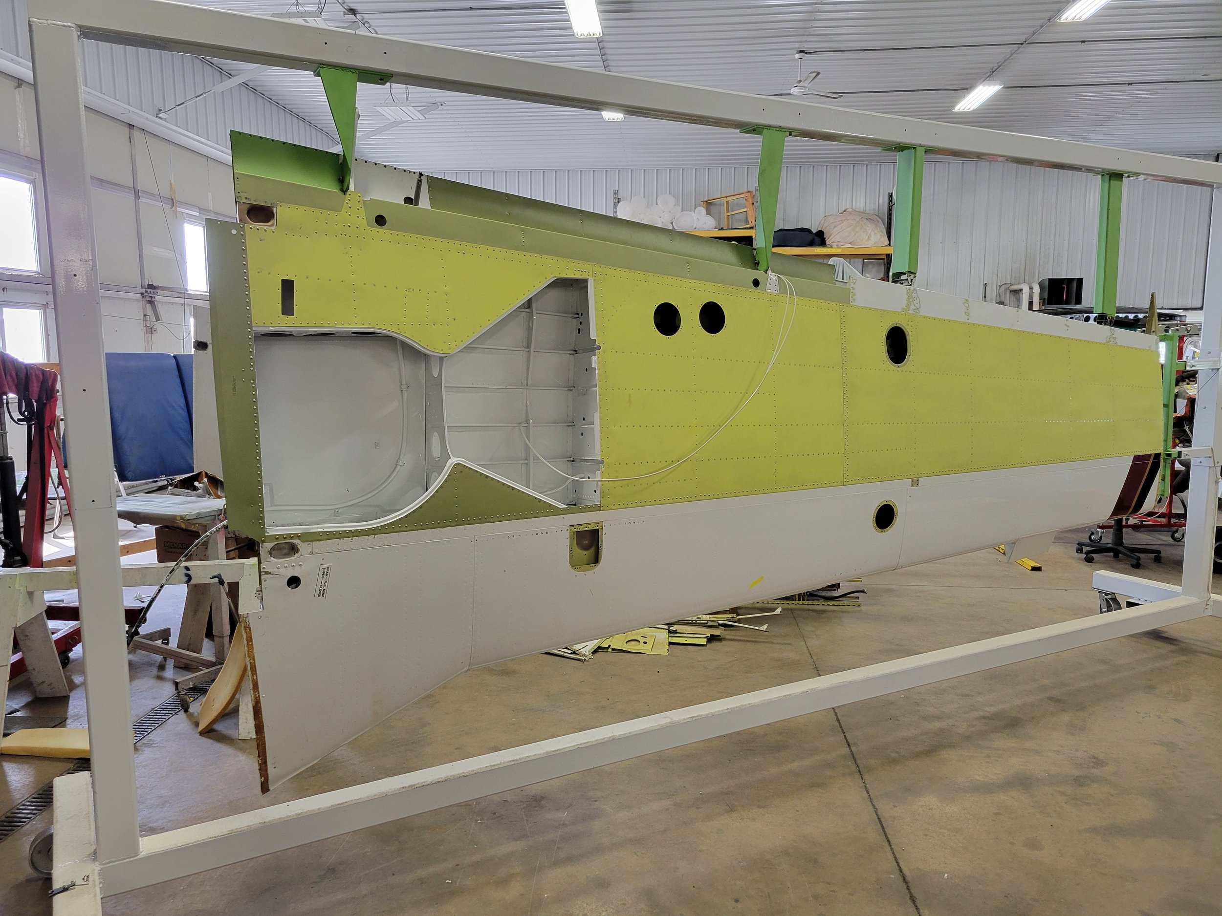 A large aircraft fuselage in an industrial workshop with white walls and ceiling. The fuselage is partially assembled with green and yellow panels and circular openings. Shelves with equipment and materials are visible in the background.