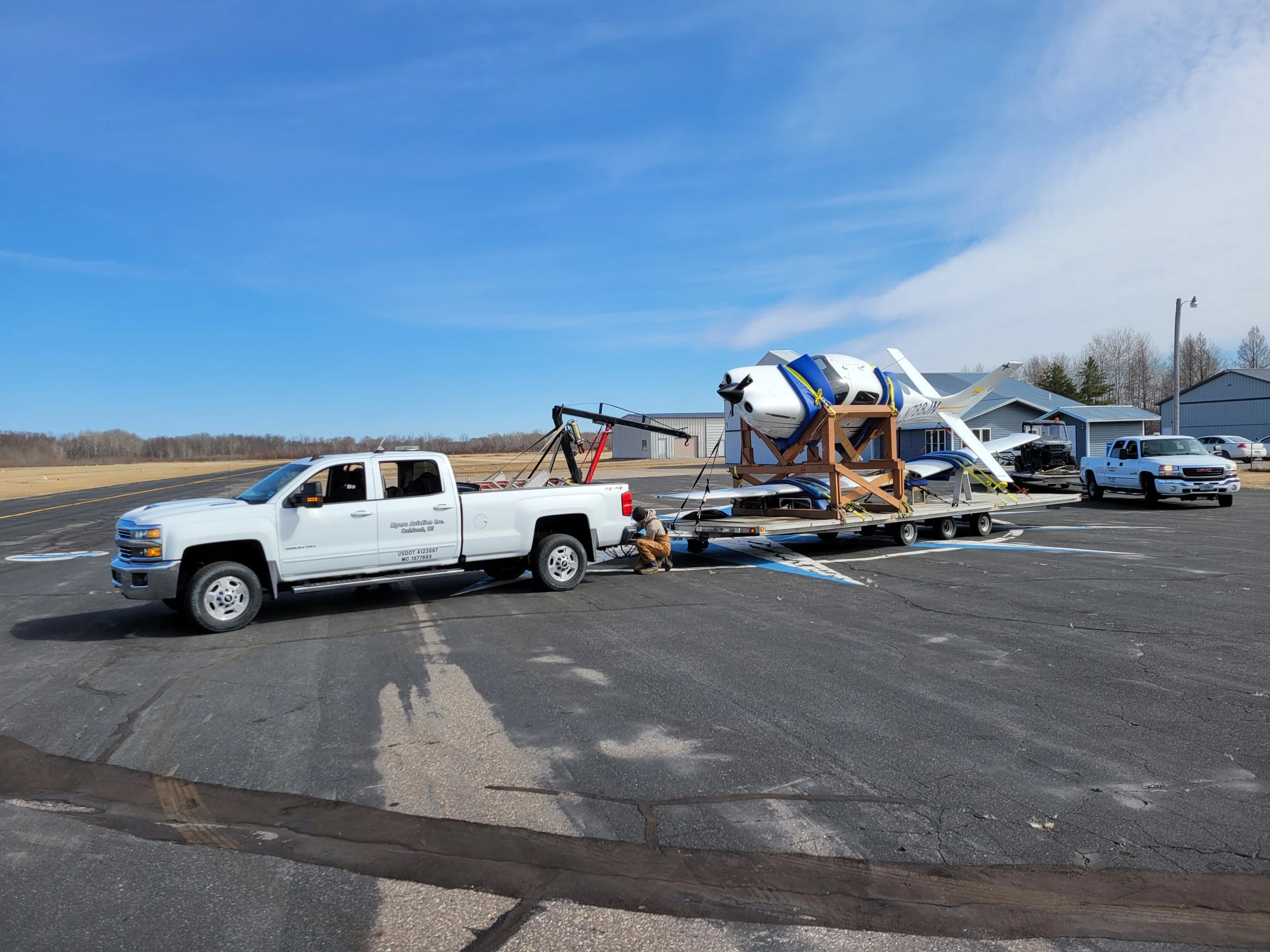 A white pickup truck carrying a small airplane on a flatbed trailer. The trailer is parked on an airport tarmac near a hangar, with a person kneeling beside the truck. The sky is clear with some clouds, and there are other vehicles and buildings in the background.
