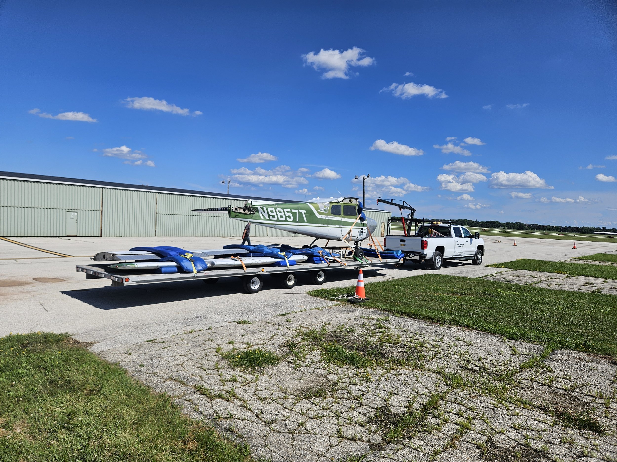 Small airplane being prepared for takeoff at an airport, with a trailer carrying equipment, a pickup truck, and clear blue skies with a few clouds.