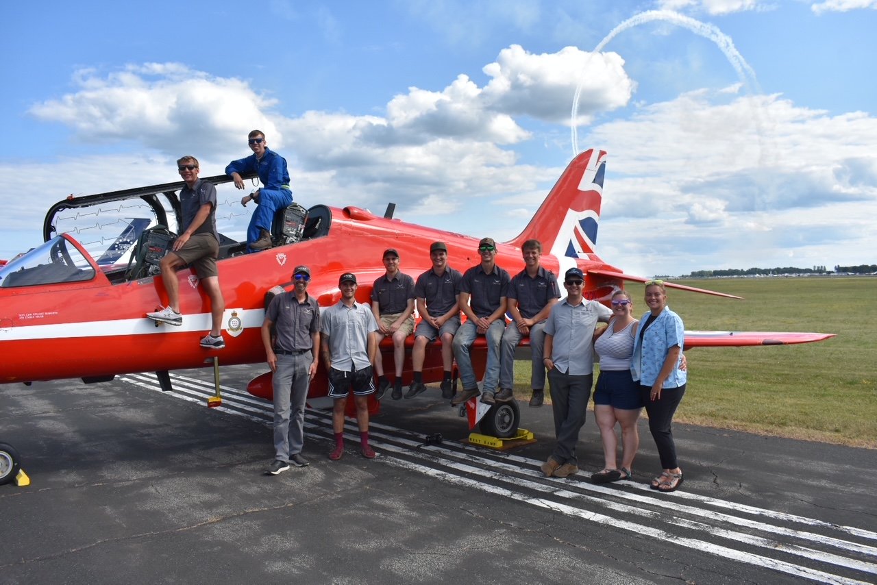 Group of people posing in front of a red fighter jet on an airfield, with some sitting on the jet's wing and others standing on the ground. The sky is partly cloudy.