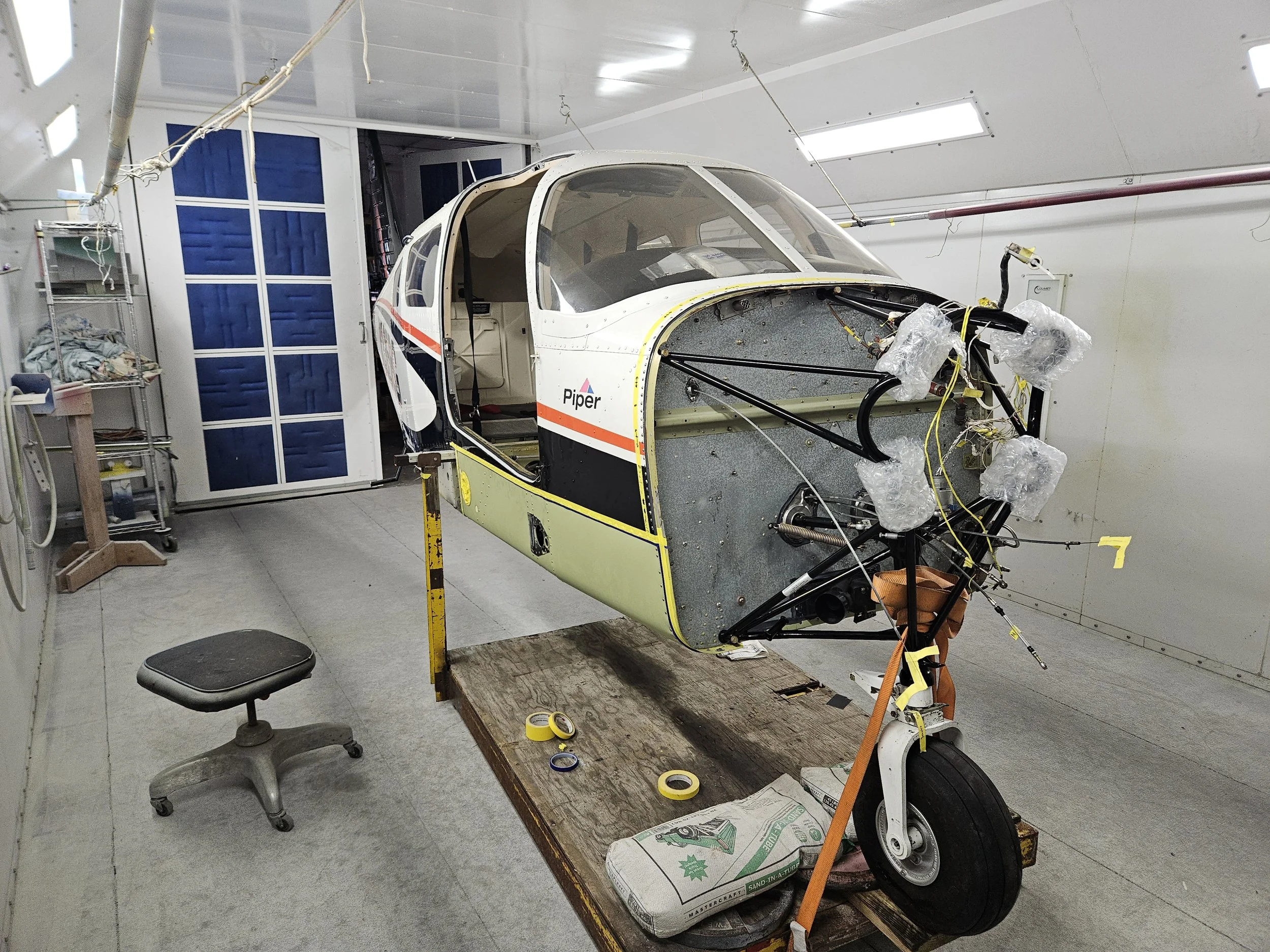 An aircraft cockpit under construction in a workshop. The fuselage is partially assembled with wires and components visible. There are tools and supplies around, including a wooden worktable with yellow tape, a stool, and a sealed bag of sand in front of the aircraft.