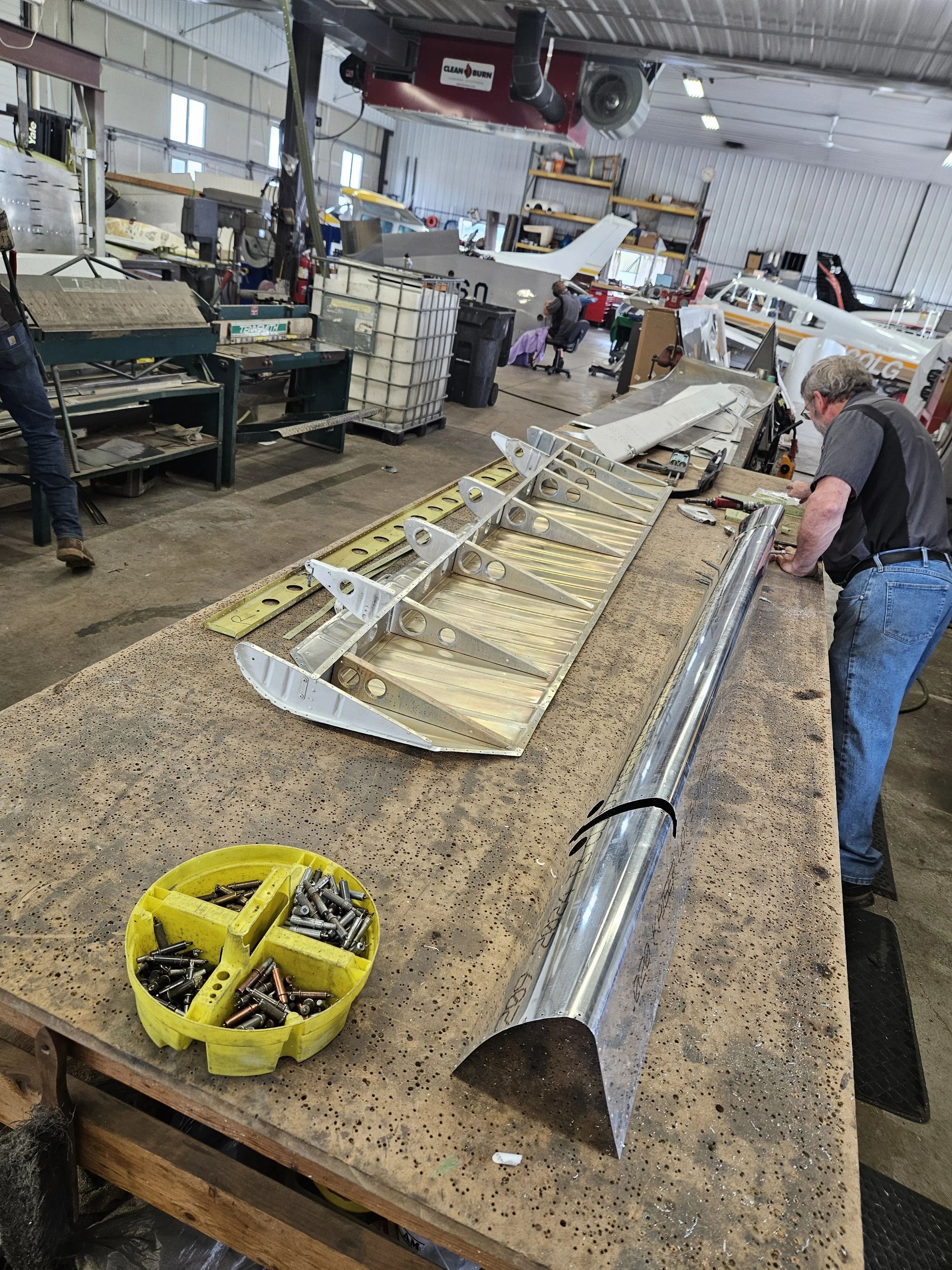 A workshop with airplane parts and tools on a workbench, including a metal fuselage section, screw bins, and workers assembling or repairing aircraft structures.