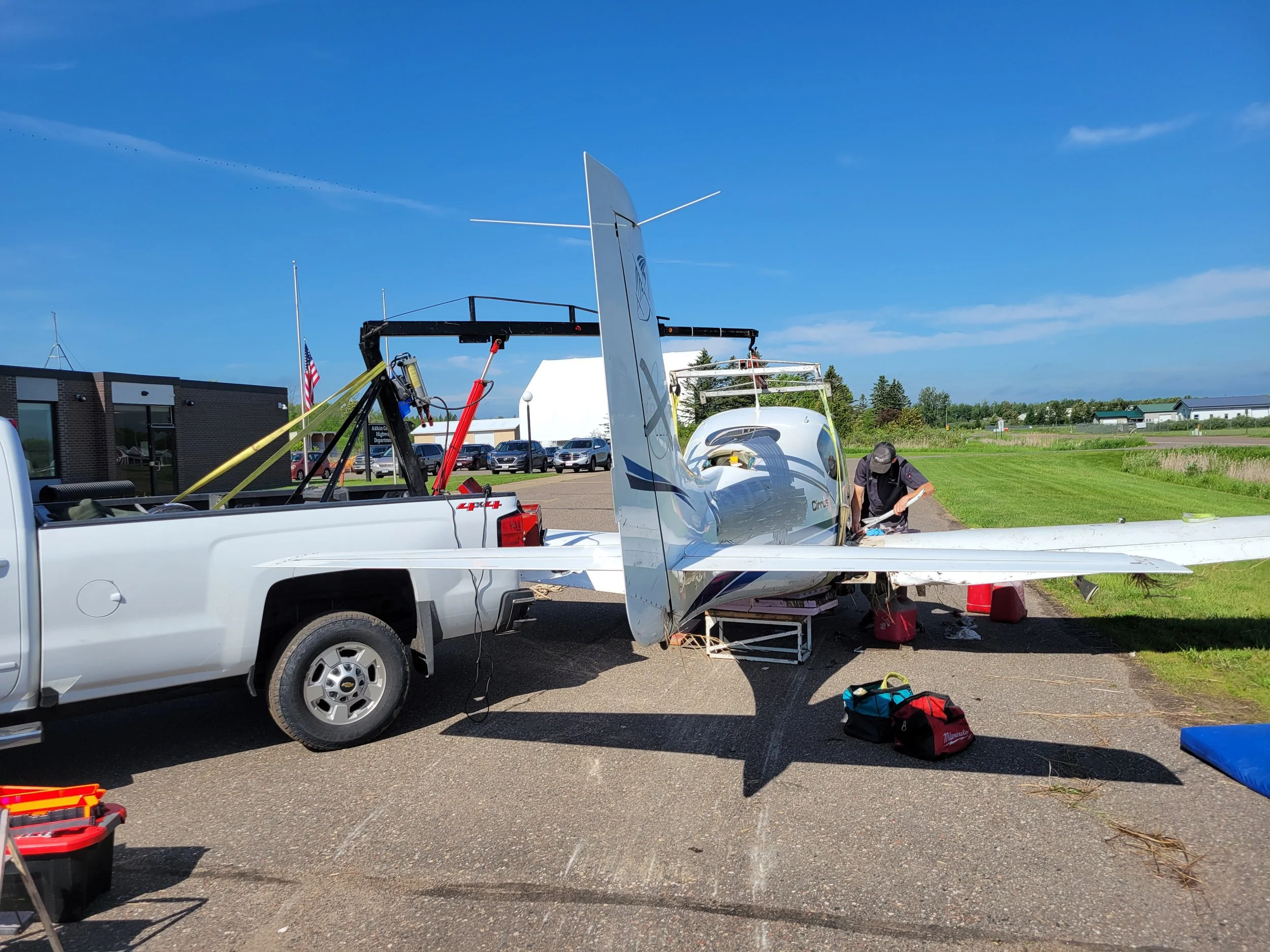 A person working on a small aircraft outside in a parking lot, with tools and equipment around, and a building and trees in the background.