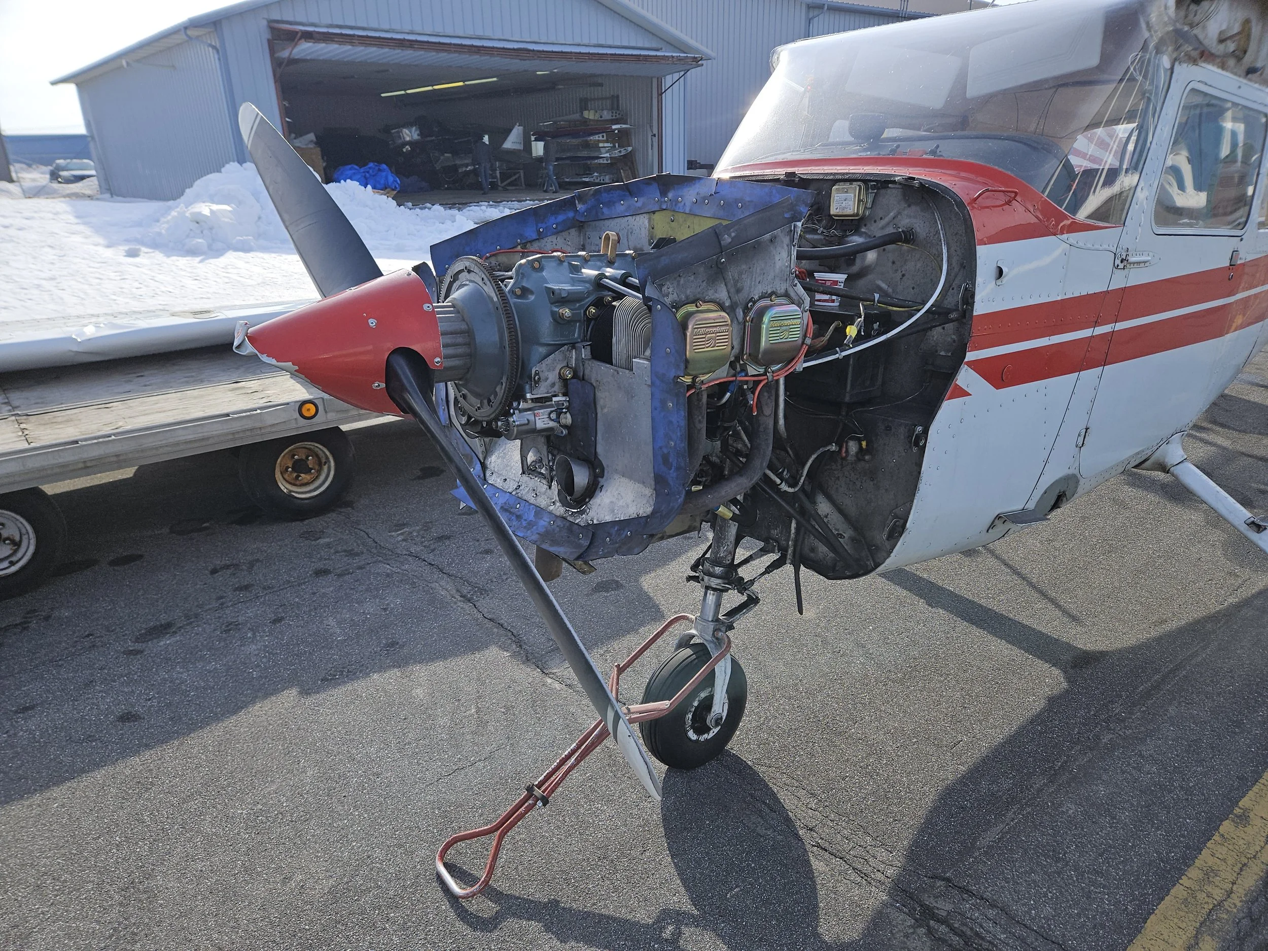 A small airplane with its engine cowling removed, showing the engine inside, parked on snow-covered ground near a hangar.