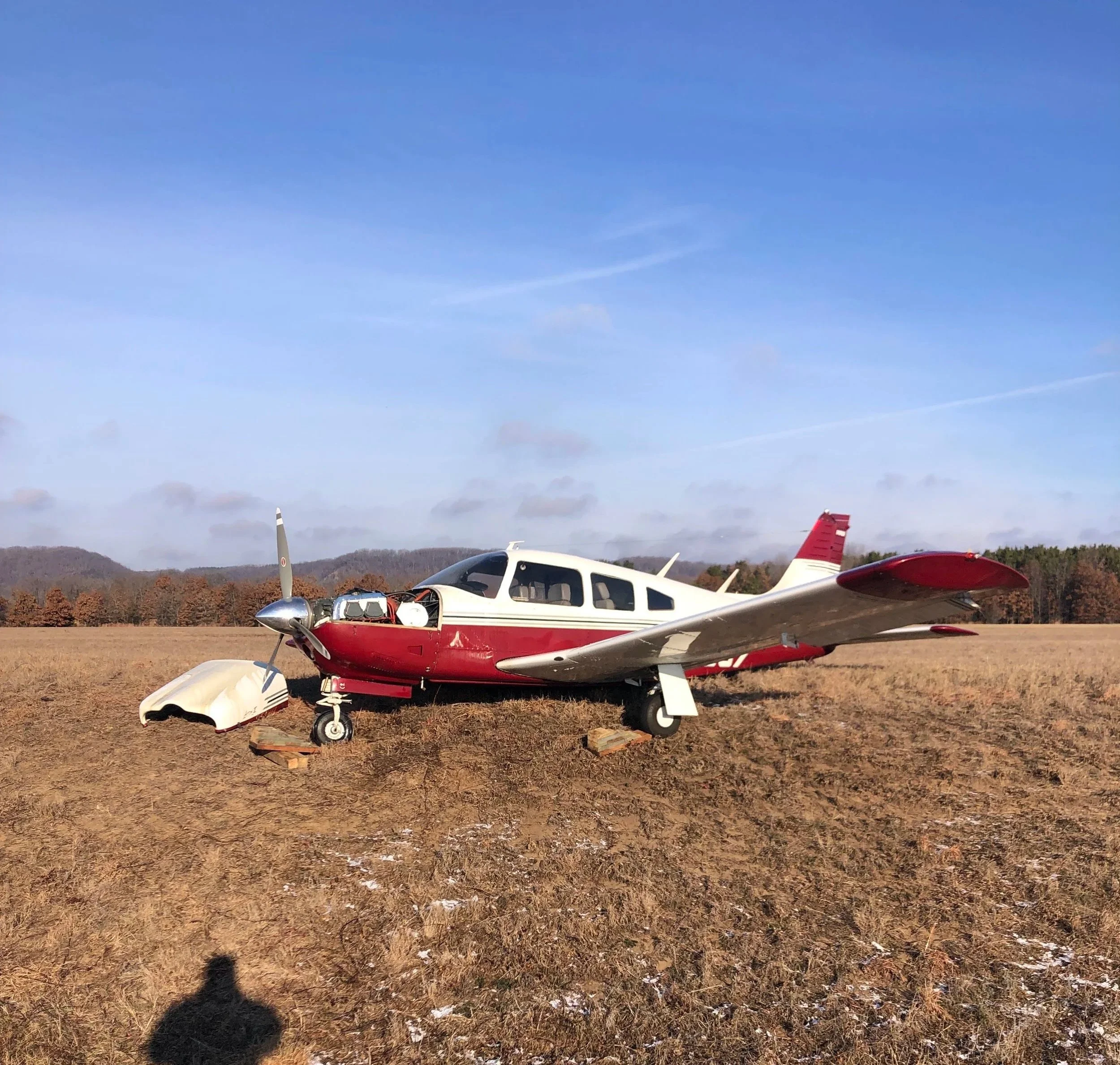 Small airplane on a dry grassy field with trees and hills in the background under a blue sky.