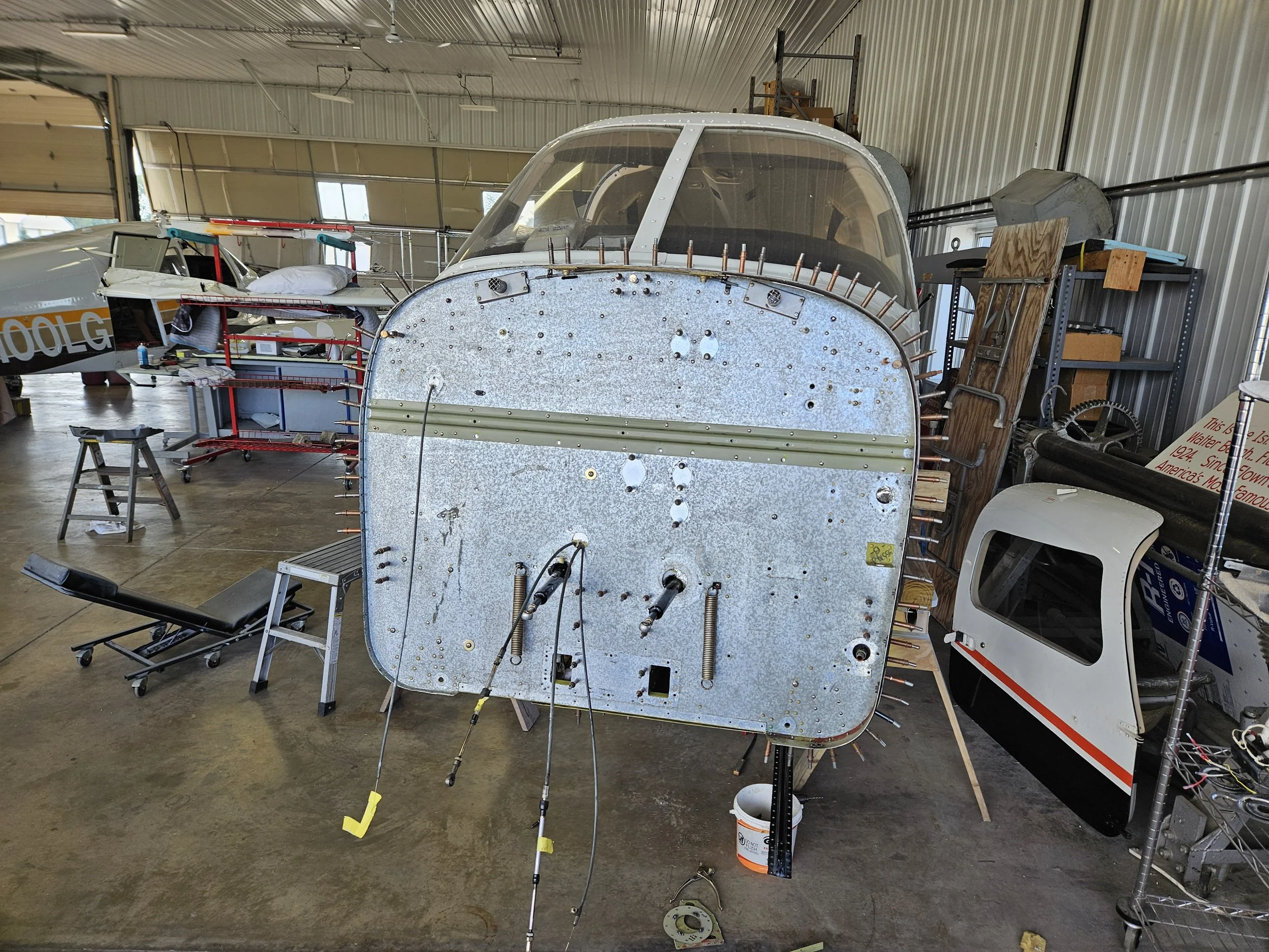 Image shows the interior of an aircraft hangar with a partially assembled vintage plane fuselage, surrounded by tools and equipment, including a workbench, a ladder, and various parts.