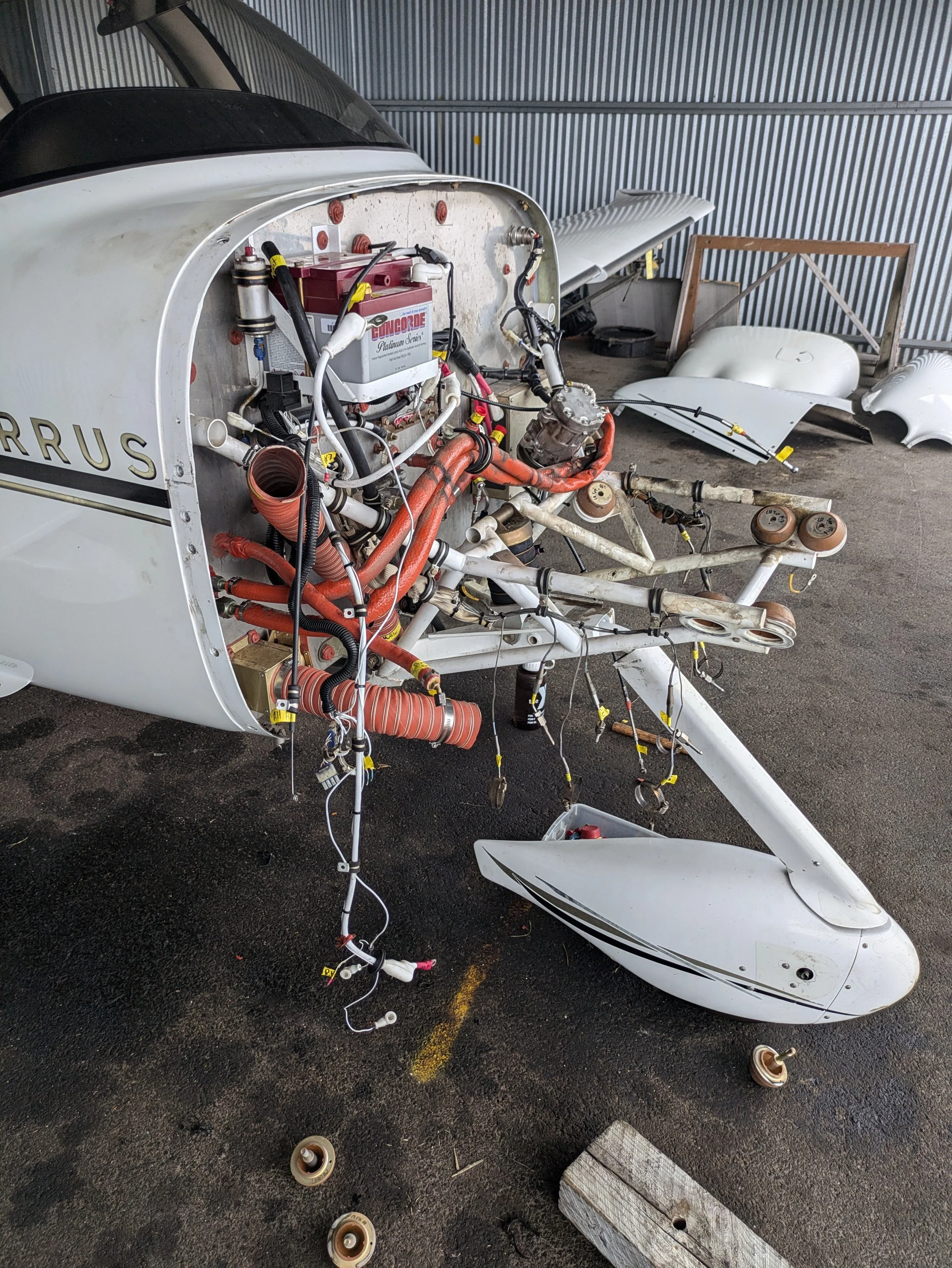 Partially assembled small aircraft fuselage with exposed internal wiring and components, white body panels, and various tools and parts on the ground, in a hangar with corrugated metal walls.