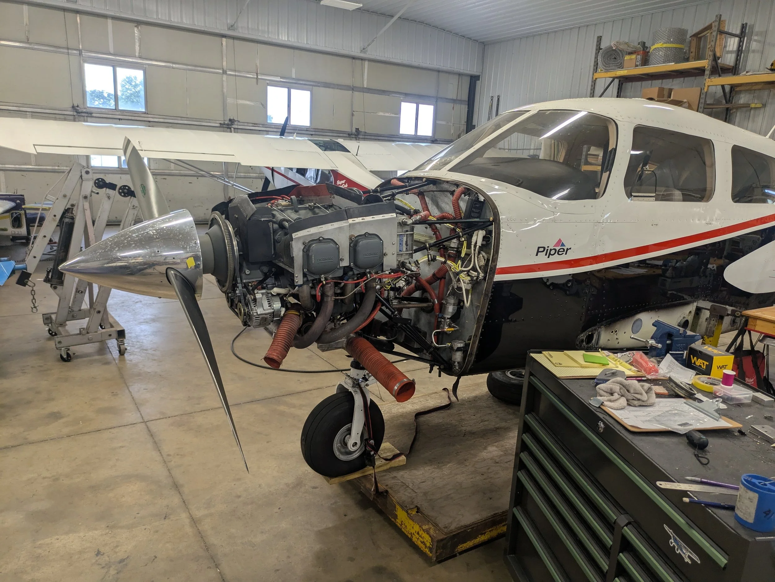 Inside an aircraft hangar, a white small aircraft with an open nose and exposed engine components is under maintenance. Tools and equipment are scattered around, and there are shelves with supplies in the background.
