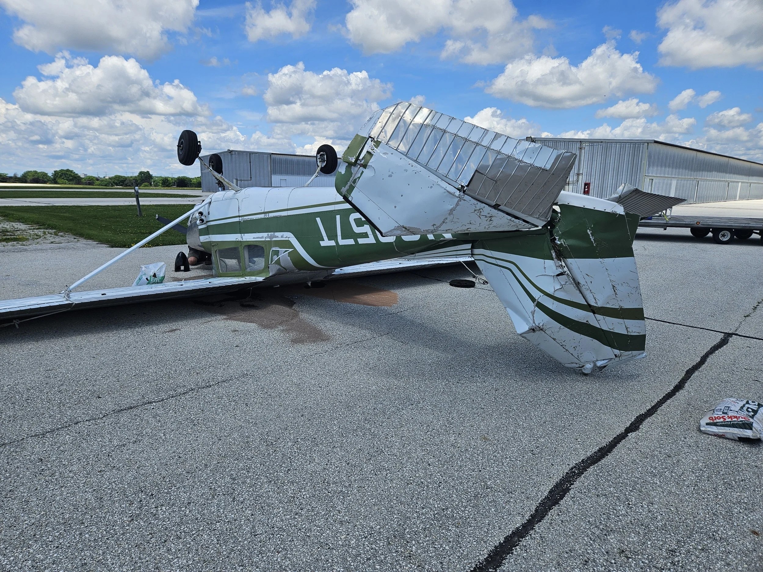A small airplane has crashed and flipped onto its side on an airport tarmac, with visible damage to the fuselage and wings, under a partly cloudy sky.