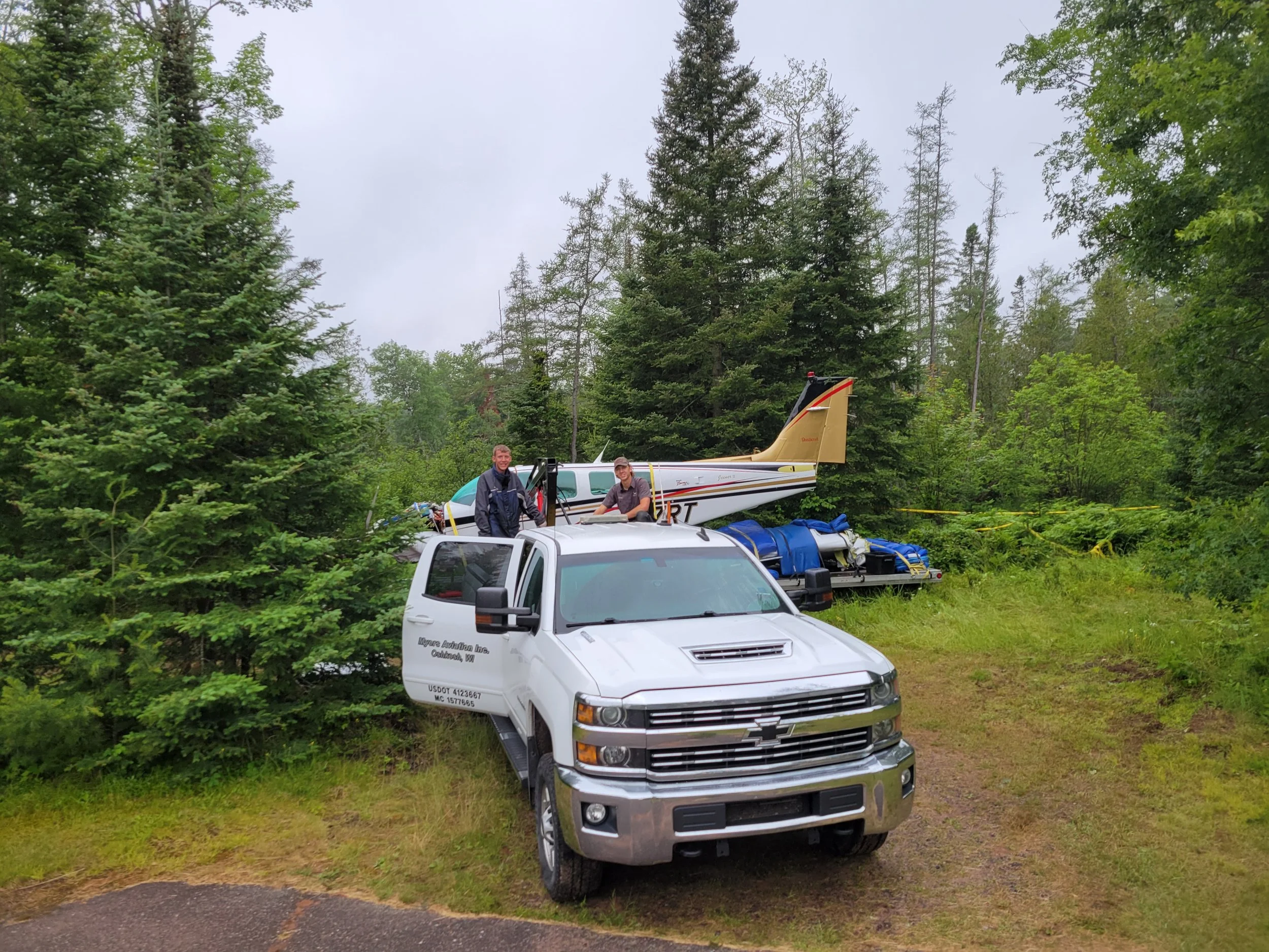 Two men stand next to a small airplane and a white truck in a forested area with tall green trees and overcast sky.