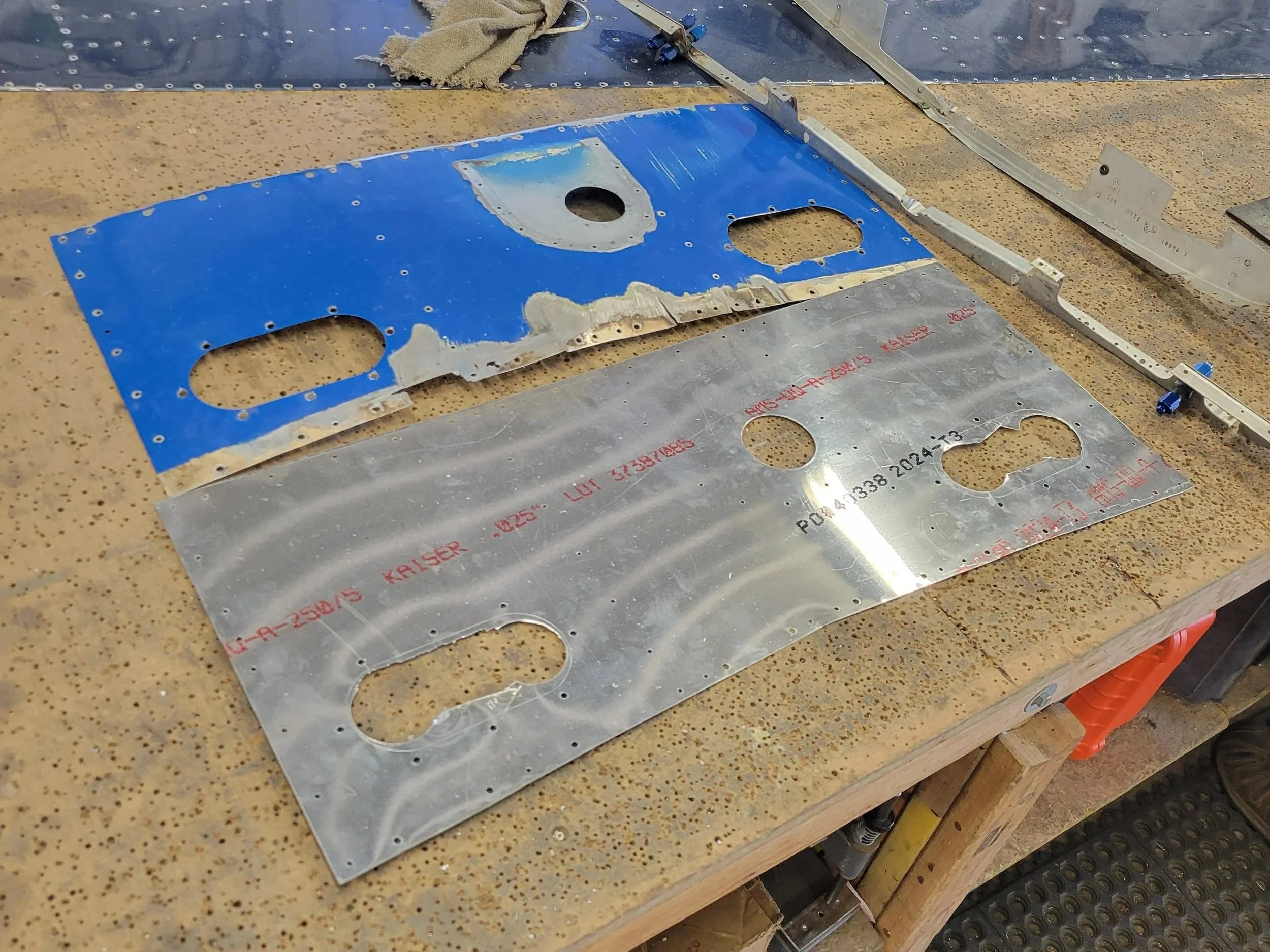 Close-up of aircraft parts on a workbench, including metal sheets with drilled holes, blue protective tape, and measuring tools used for assembly or repair.