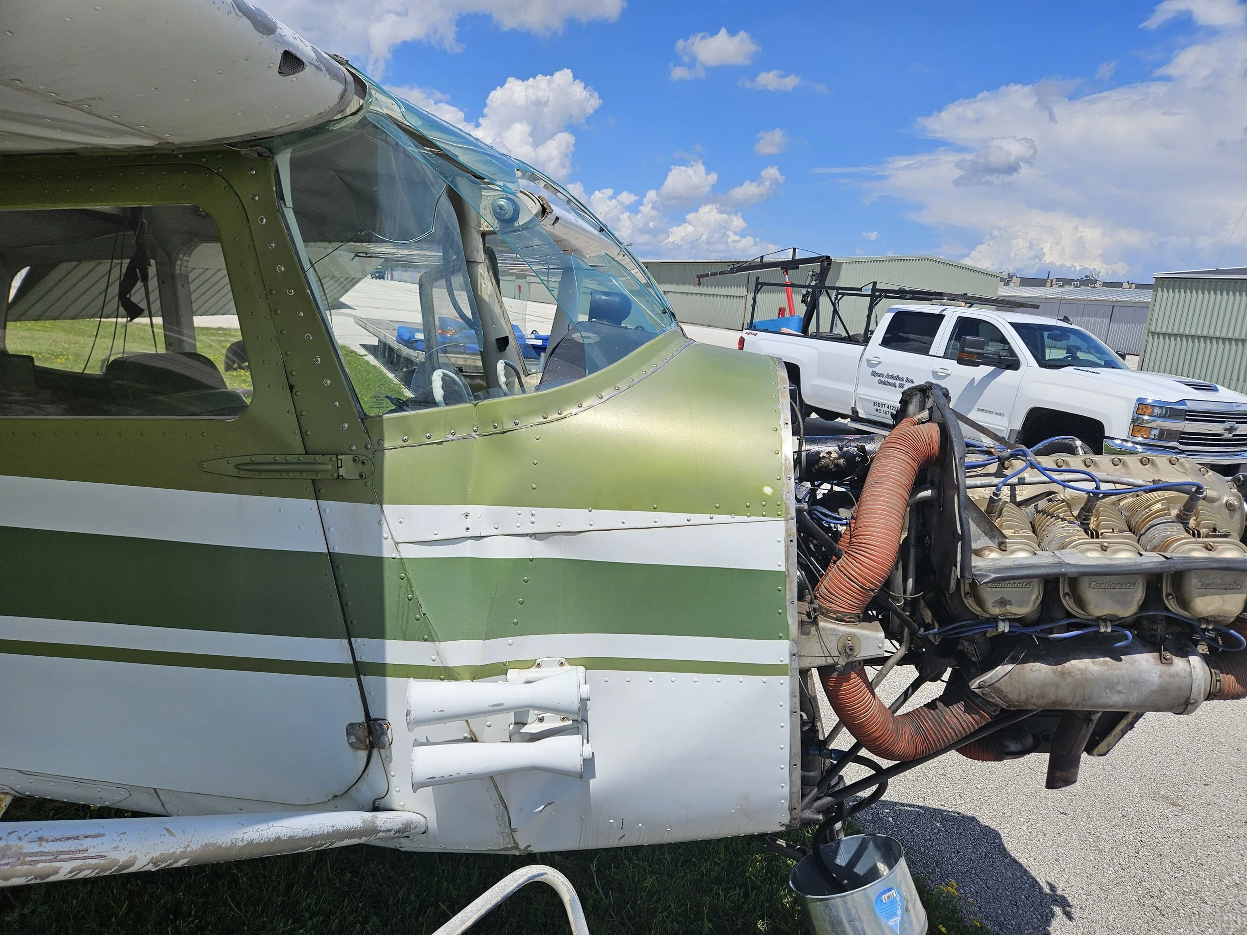 The image shows an aircraft's cockpit and engine with some parts exposed, situated outdoors next to a white truck with text on the door, on a cement surface with a blue sky and clouds overhead.