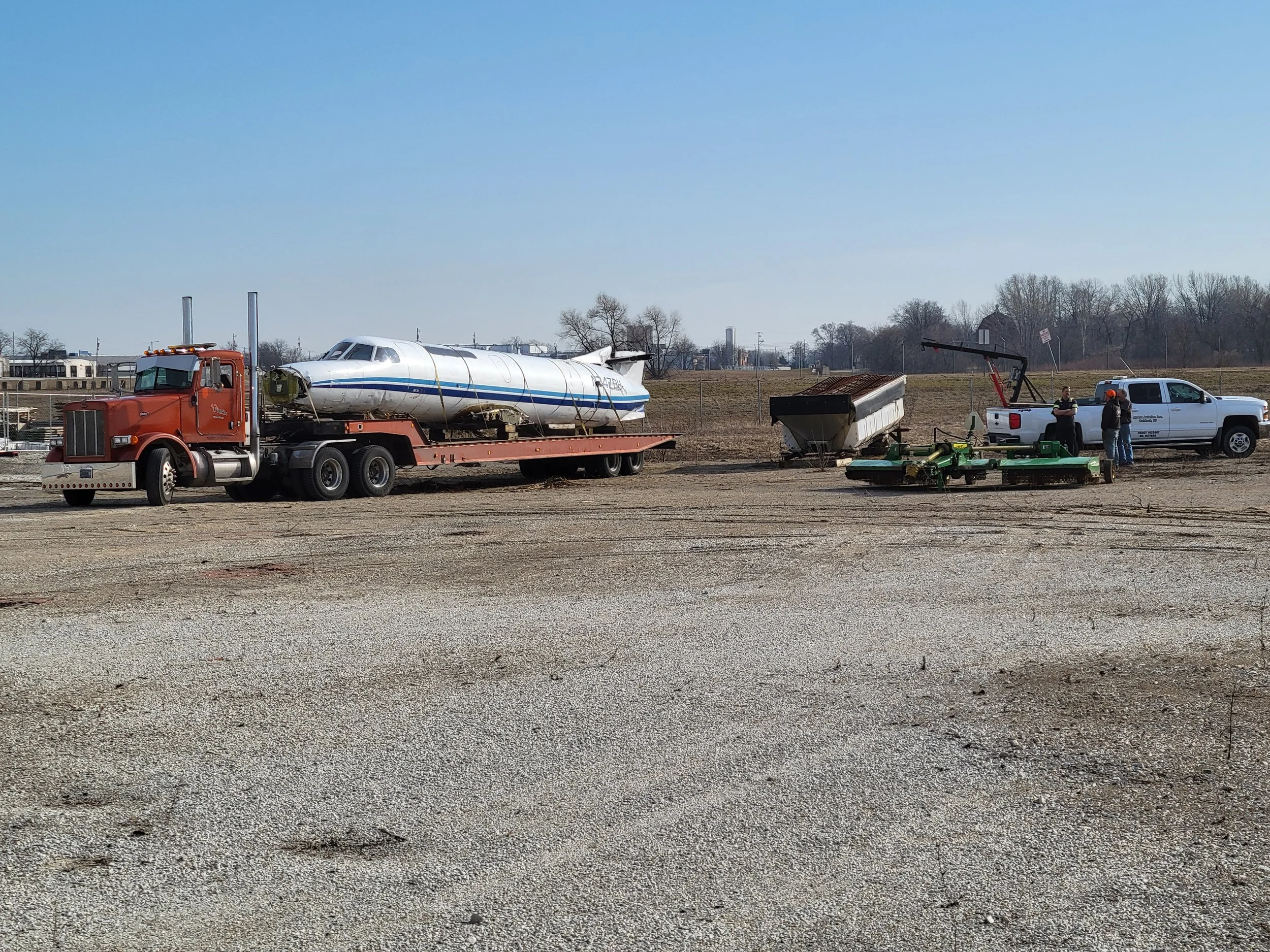 A red semi-truck transporting an aircraft fuselage on a trailer, with two people and farming equipment nearby, on a dirt field under a clear blue sky.