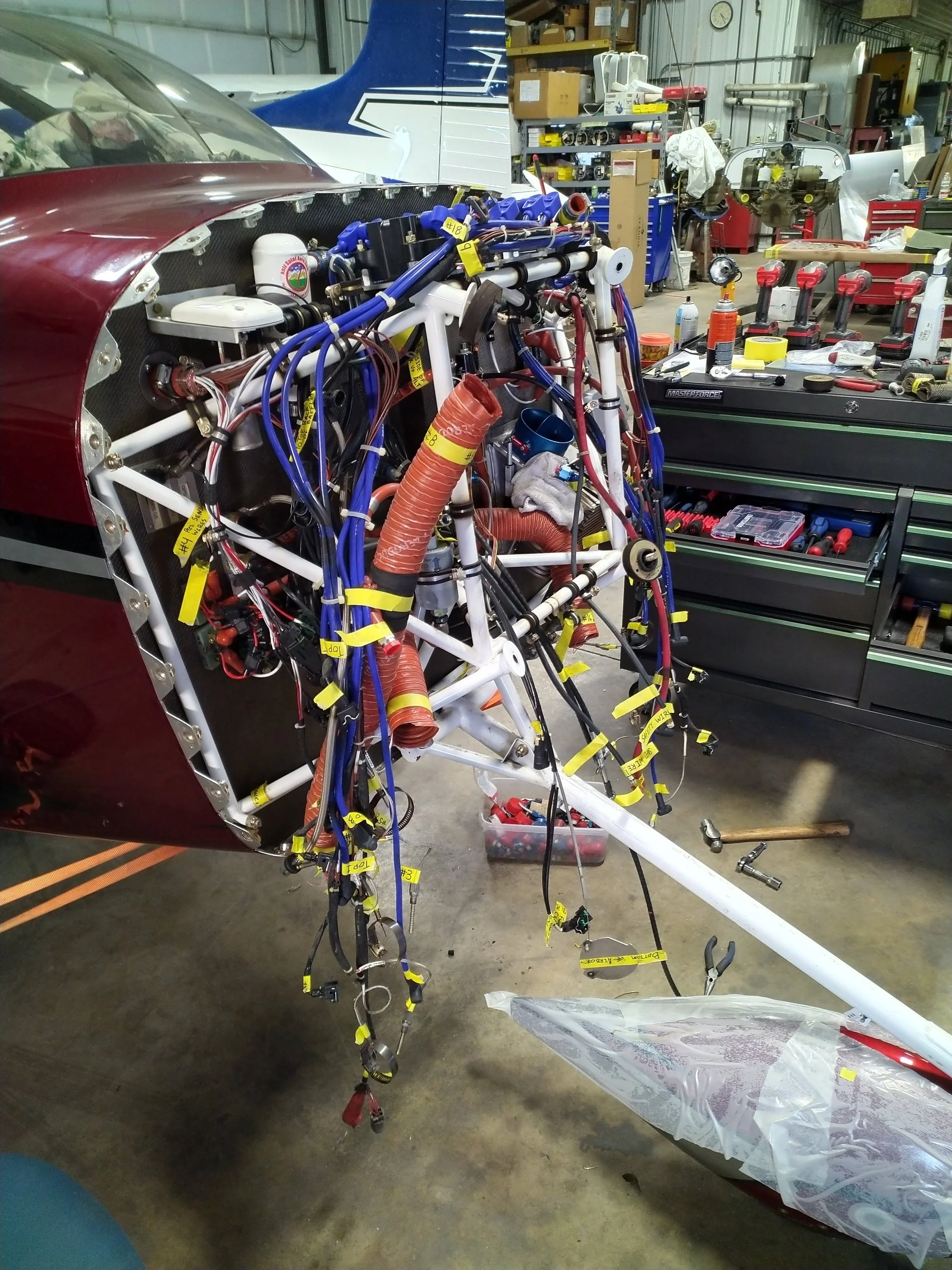 Inside an aircraft hangar, a partially assembled light aircraft with exposed wiring, tubing, and structural components. The workspace is cluttered with tools, toolboxes, and various aircraft parts.