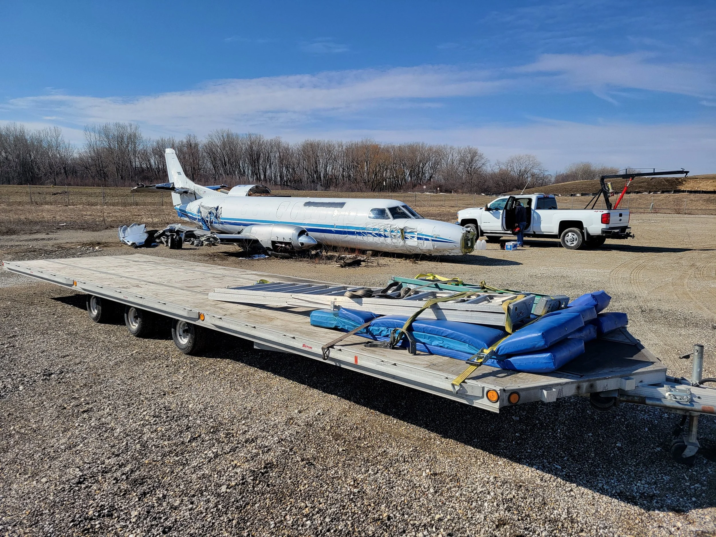Debris of an airplane, including broken fuselage and parts, next to a white pickup truck with a camera crane on the bed, in an open field with a fence and trees in the background.