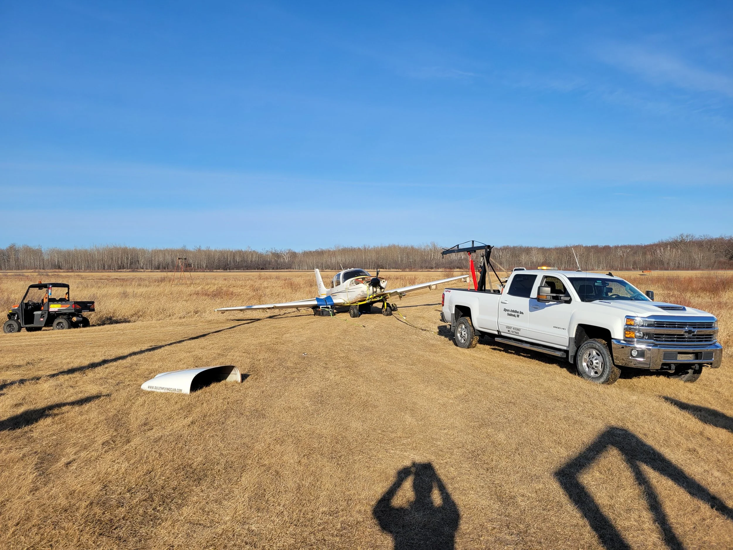 A small aircraft on a grassy field with various vehicles and equipment around it, including a white pickup truck, a utility vehicle, and helicopter equipment, under a clear blue sky.