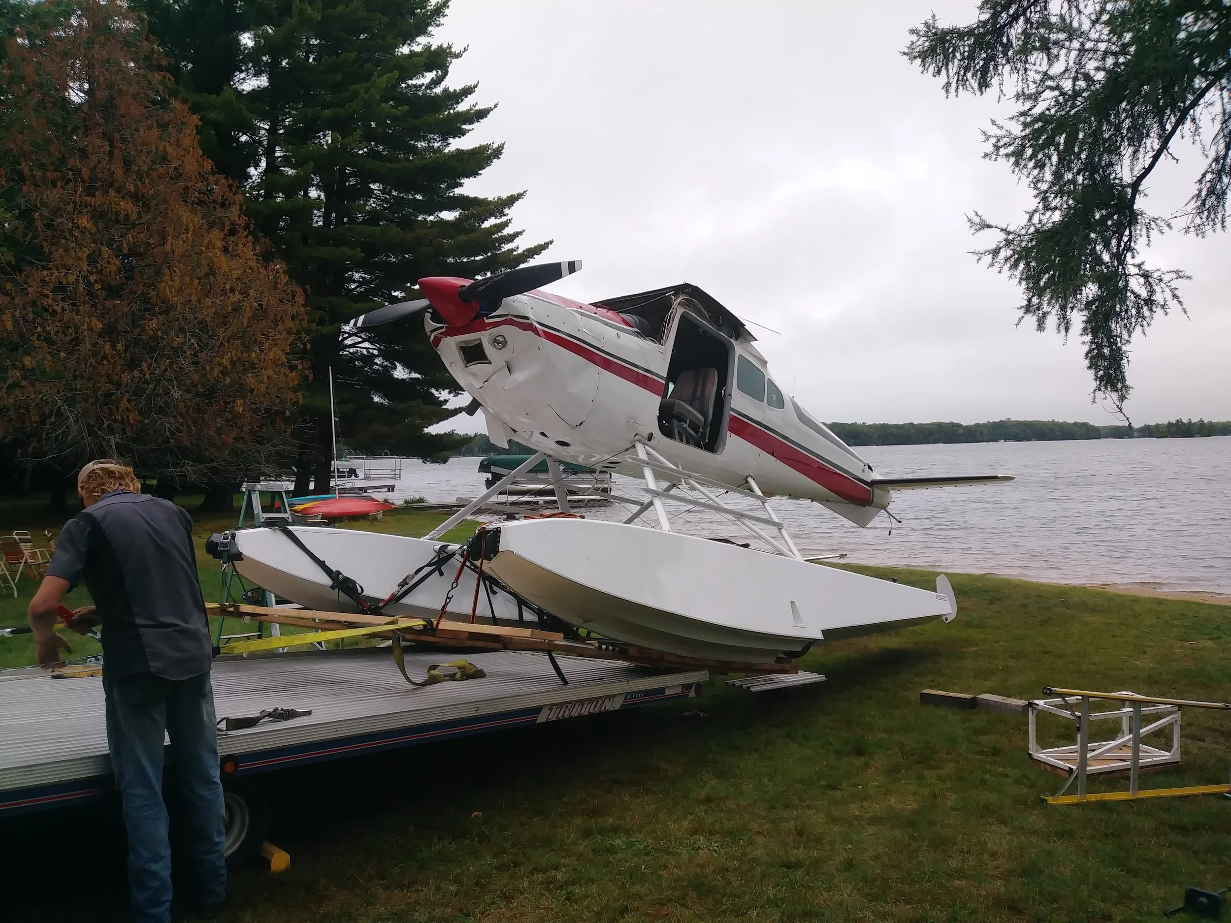 A small airplane is being loaded onto a trailer by a person near a lake, with trees and an overcast sky in the background.
