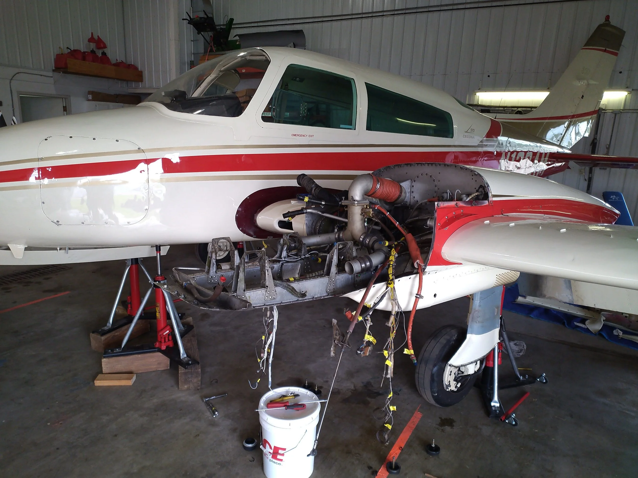 Inside a hangar, a racing aircraft is undergoing maintenance, with parts and wiring exposed, and tools on the floor.