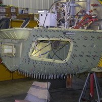 A large aircraft fuselage section under construction in an industrial setting, with visible windows and interior framing.