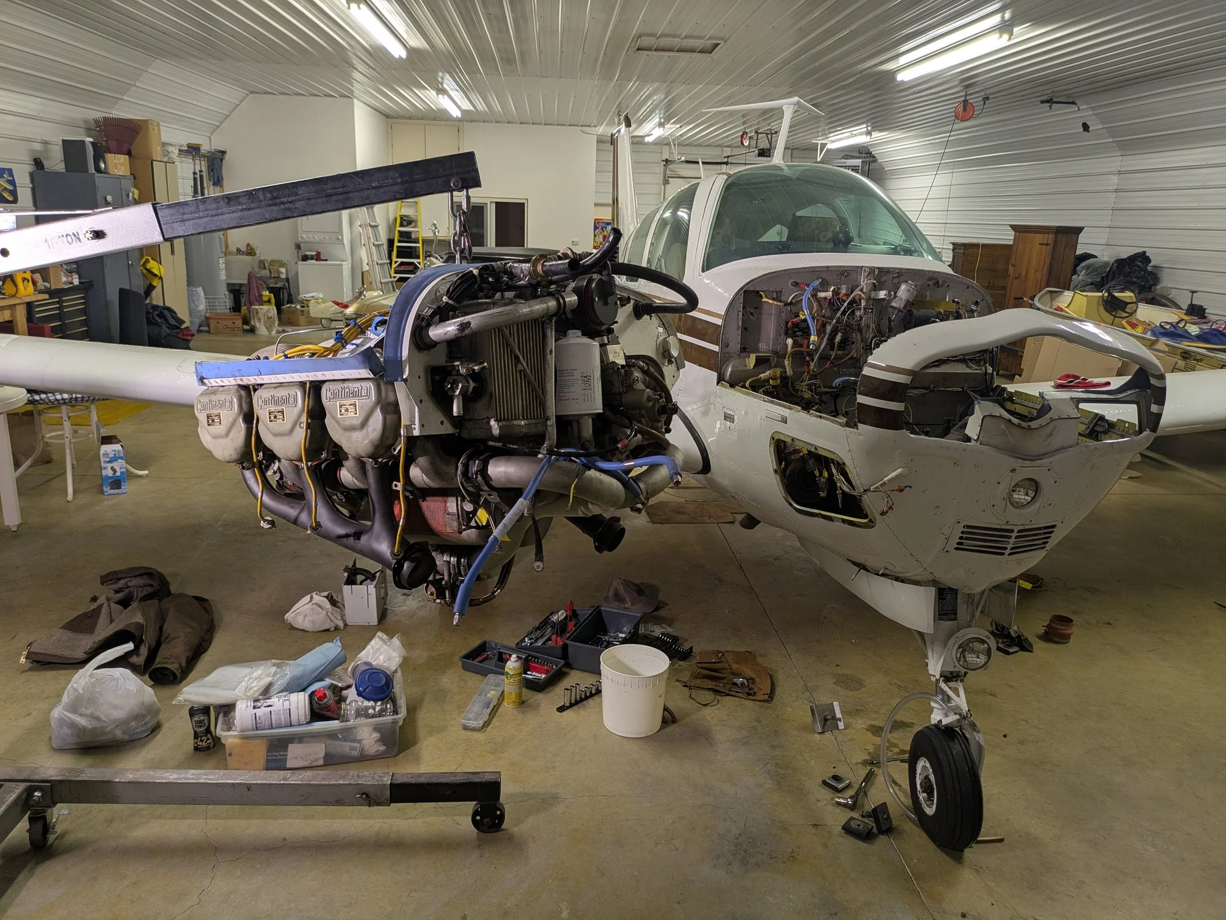 The image shows a garage with an airplane under construction. The airplane's engine and some of its cockpit are visible, with various tools and parts scattered around. The ceiling has fluorescent lights, and there are shelves with various items in the background.