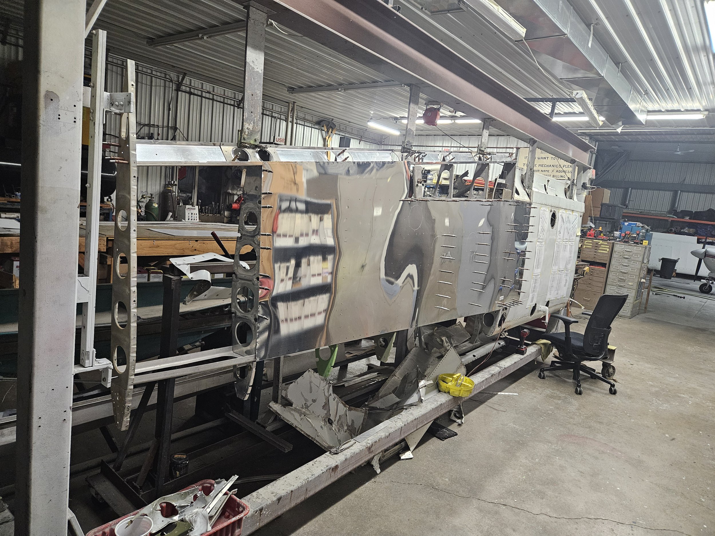 An aircraft fuselage in a workshop under construction, with a shiny metal exterior and an open section showing internal framework, surrounded by tools and equipment.