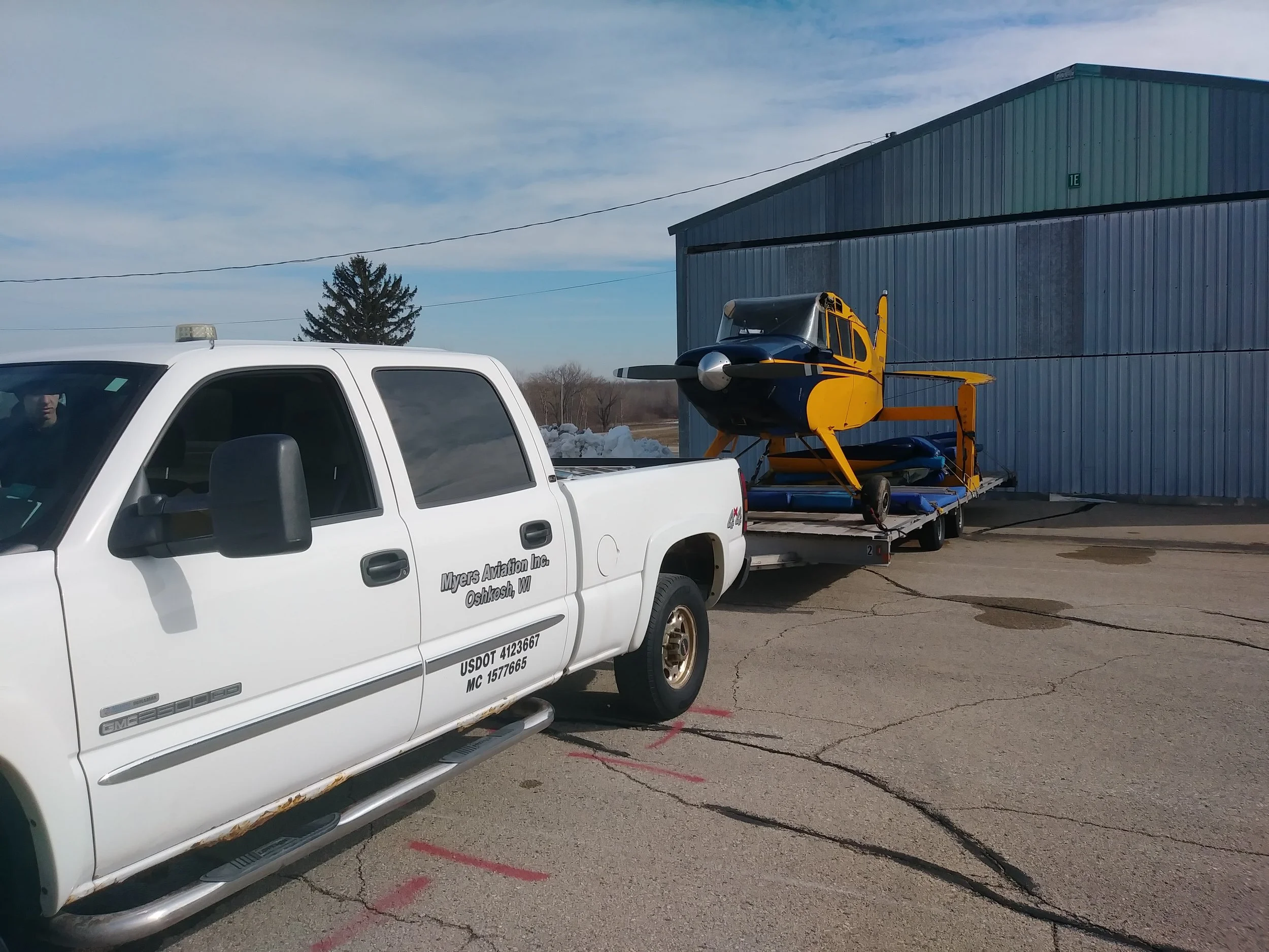 A white pickup truck with text on the door, parked on a cracked pavement, towing a trailer with a yellow and black aircraft. There is a blue industrial building in the background and a partly cloudy sky.