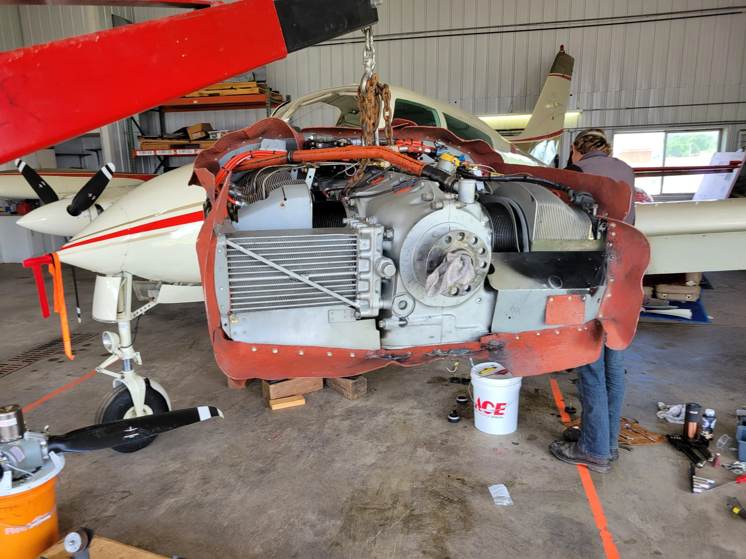A small aircraft with an exposed engine inside a workshop, with a person working on it. The aircraft has a white body with red accents and a propeller. Tools and equipment are scattered around.