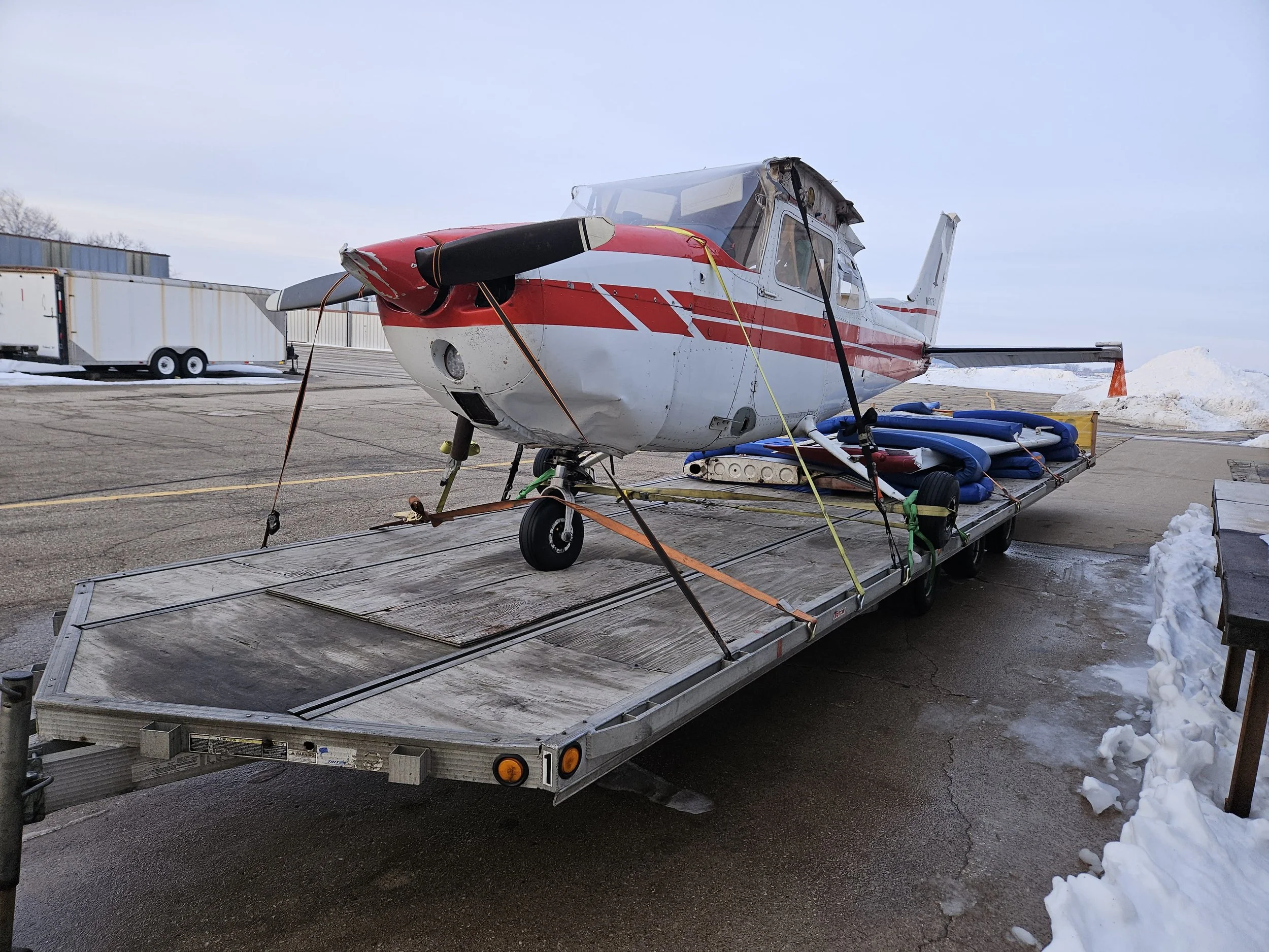 Small airplane with red and white exterior on a flatbed transport trailer, tied down with straps, with snow on the ground and a cloudy sky in the background.