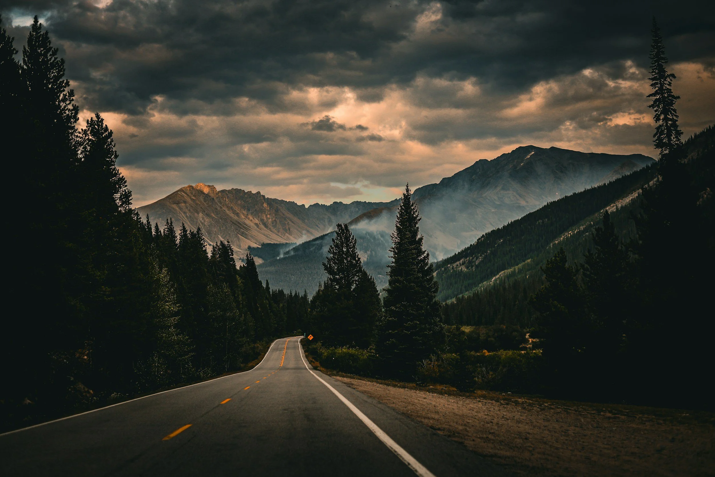 A scenic mountain road winding through dense forest with mountains in the background under a cloudy sky.