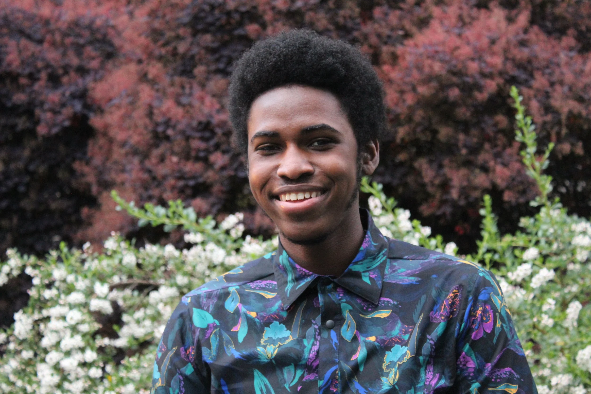 A young African American man with a broad smile, wearing a colorful patterned shirt, stands outdoors in front of blooming white flowers and dark red bushes.