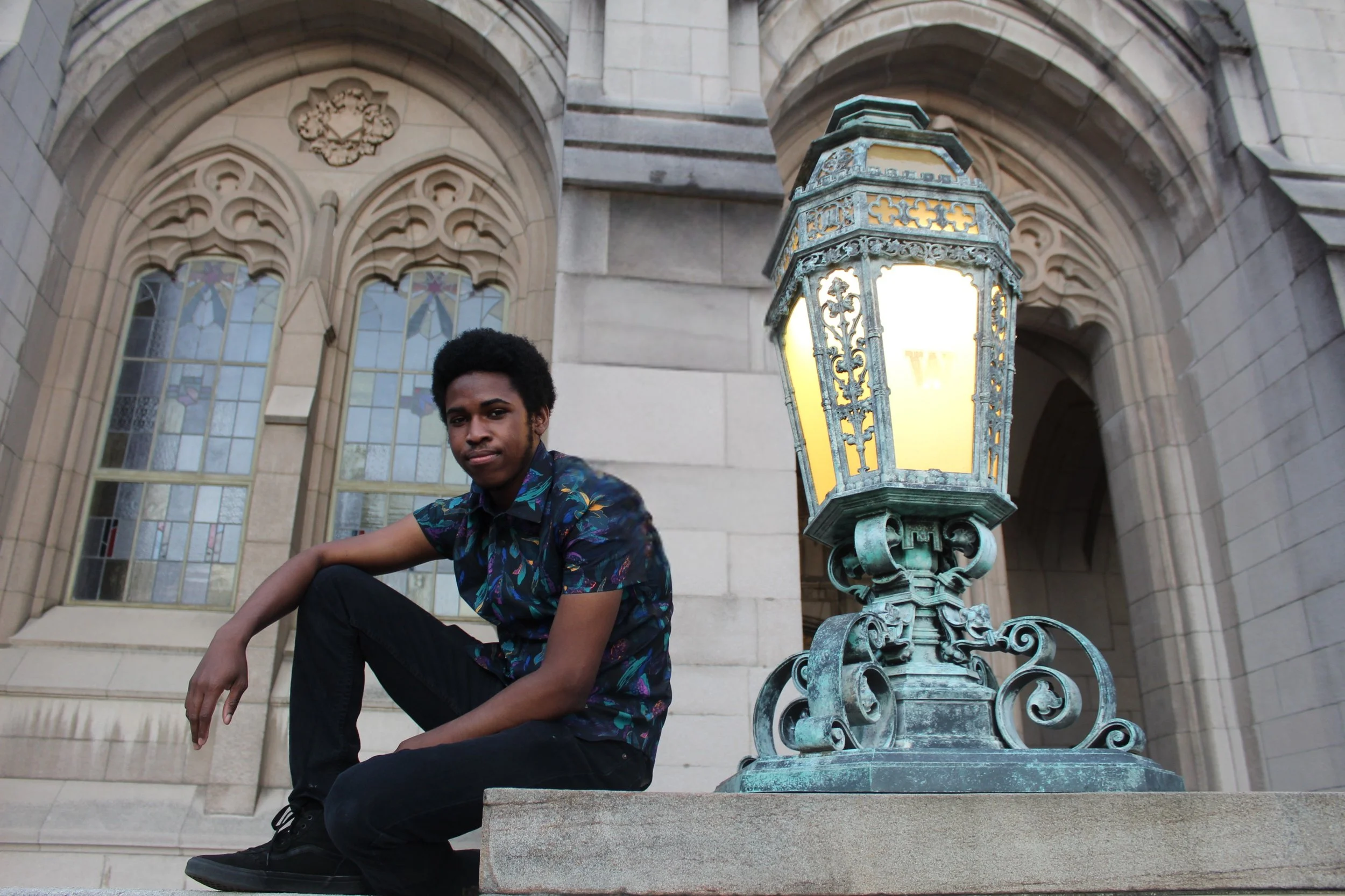 Young man with an Afro hairstyle sitting on steps outside a historic building with stained glass windows, next to an ornate street lamp.