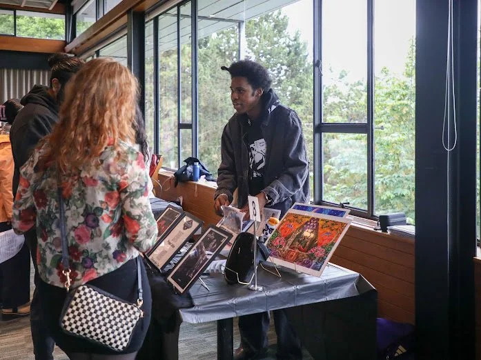 A young man at an art exhibition booth displaying colorful paintings, talking to two women, one with red hair and a floral jacket, inside a room with large windows showing greenery outside.
