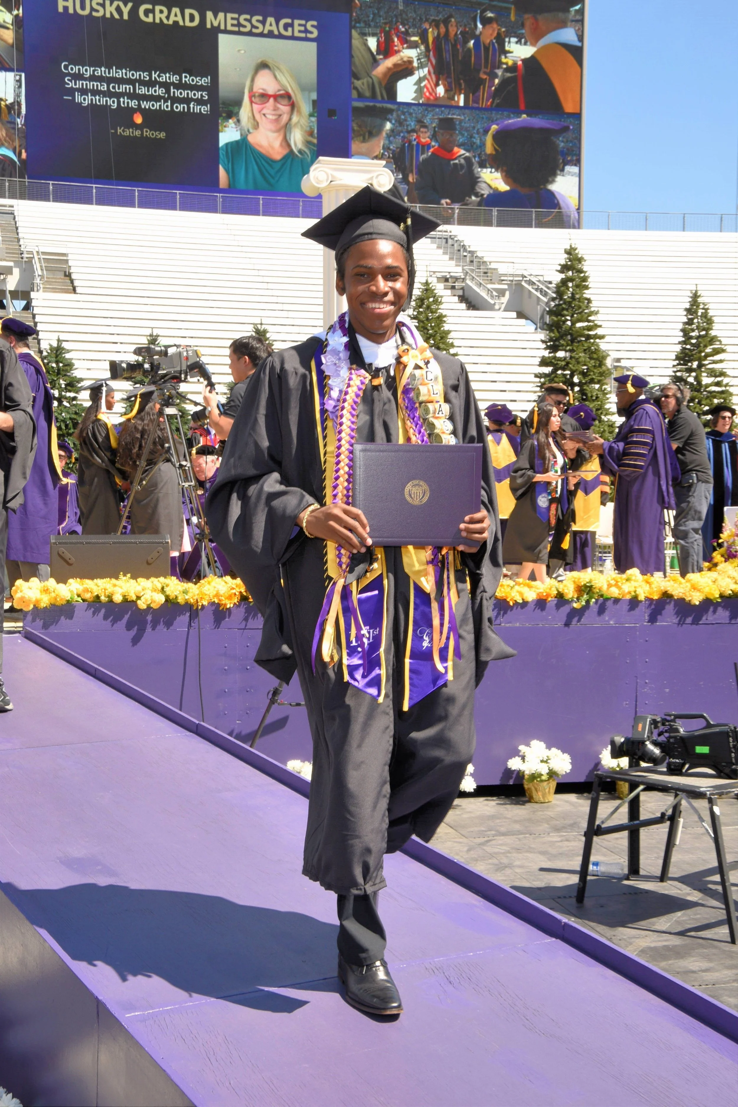 A young man wearing a black graduation cap and gown, decorated with purple, yellow, and white cords, walking on a stage at a graduation ceremony. He is smiling and holding a diploma. The background includes other graduates in purple and yellow caps and gowns, a camera crew, and a large digital screen displaying messages and images of graduates.