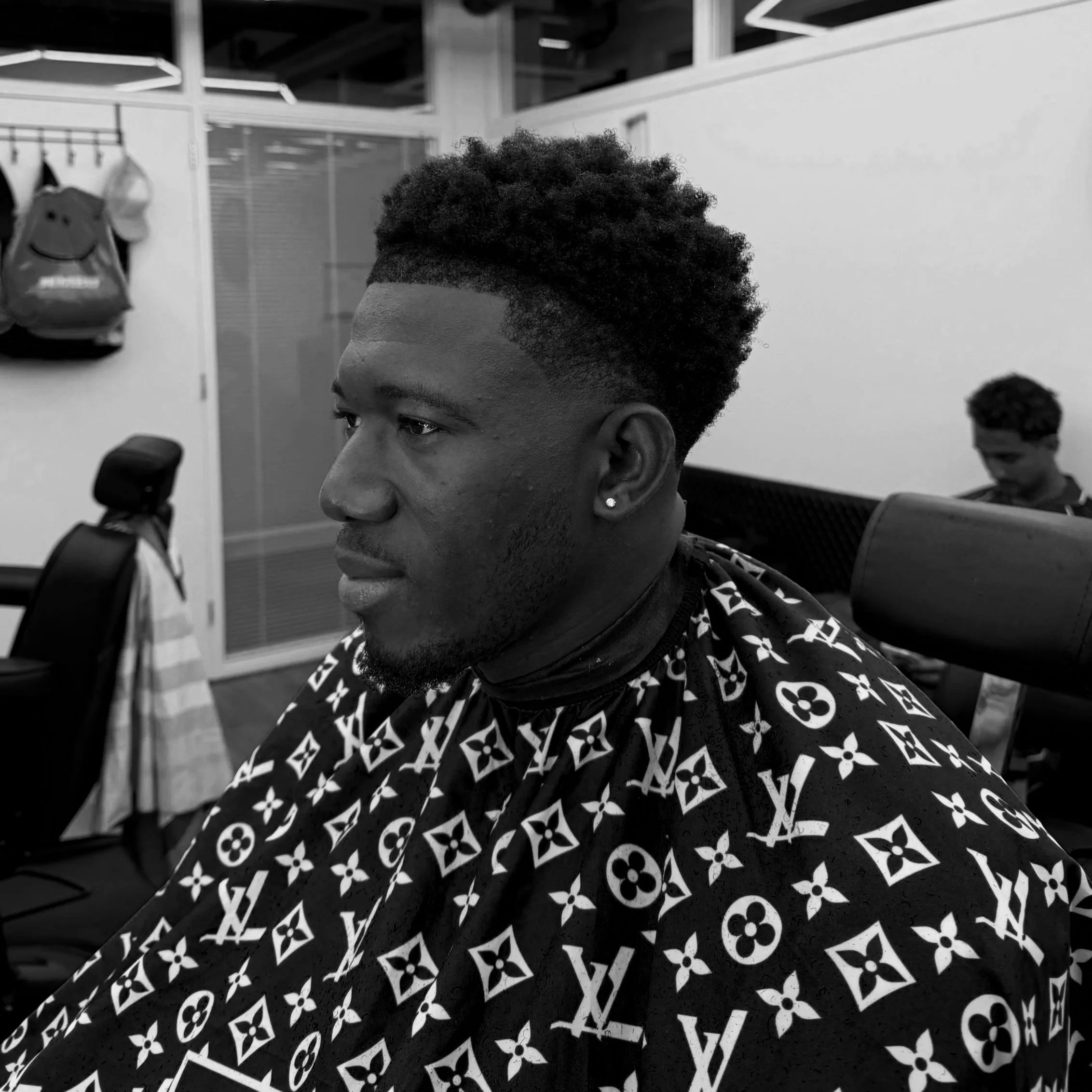 Close-up van een persoon met een getrimde, gekrulde haardos en een designer shirt, zittend in een interieurruimte, mogelijk een kapsalon of modewinkel.