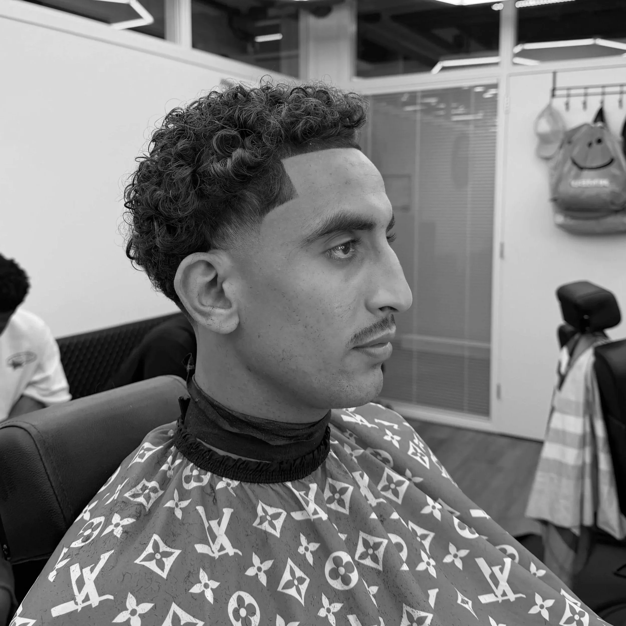 Close-up of a person's upper face, hairstyle, and eyes, seated indoors near a wall with hanging bags and a mirror.
