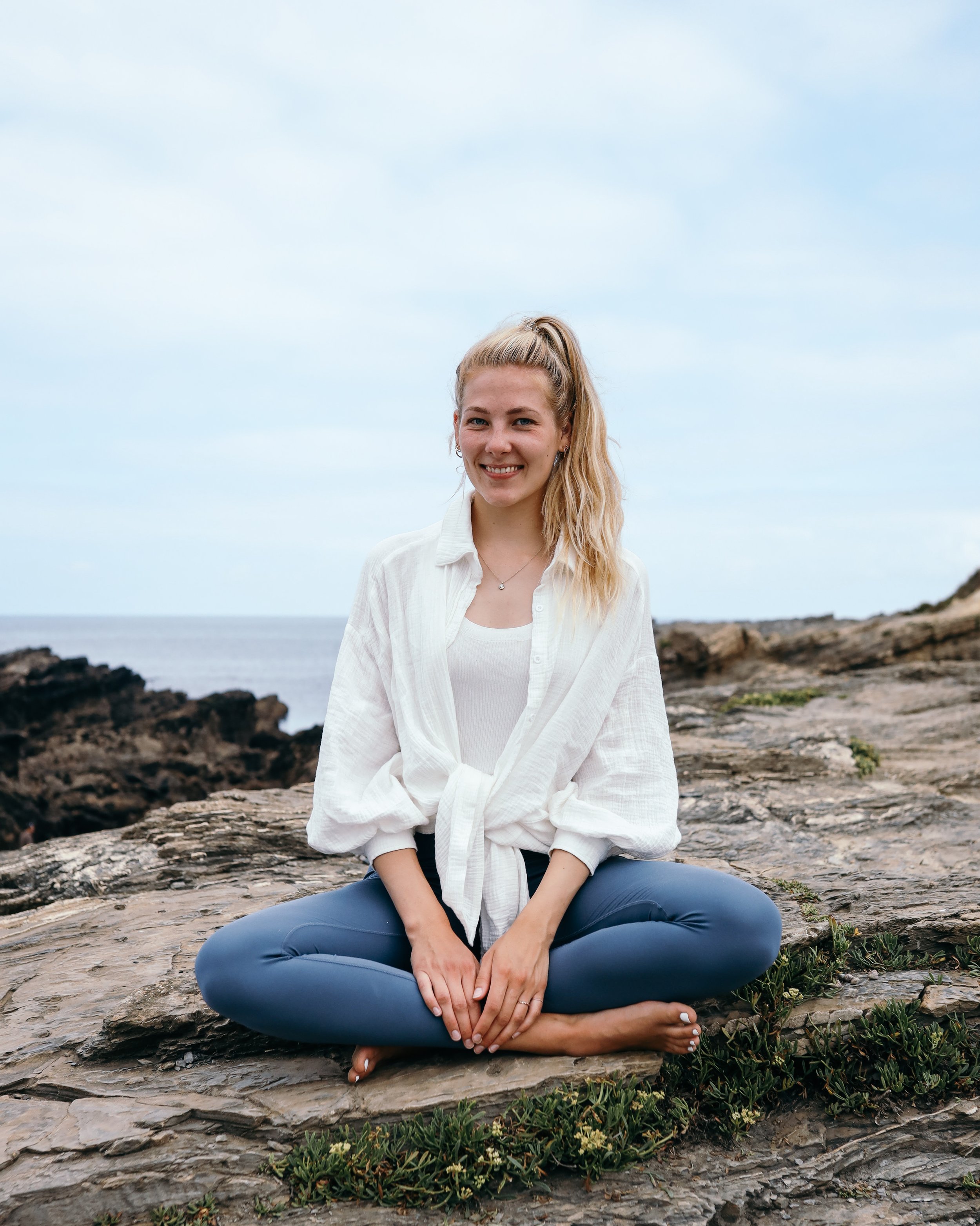 Eine junge Frau sitzt kreuzbein auf einem Felsen am Meer, trägt ein weißes Hemd und blaue Hosen, lächelt und hat blondes, langes Haar. Im Hintergrund ist der Himmel und das Meer sichtbar.