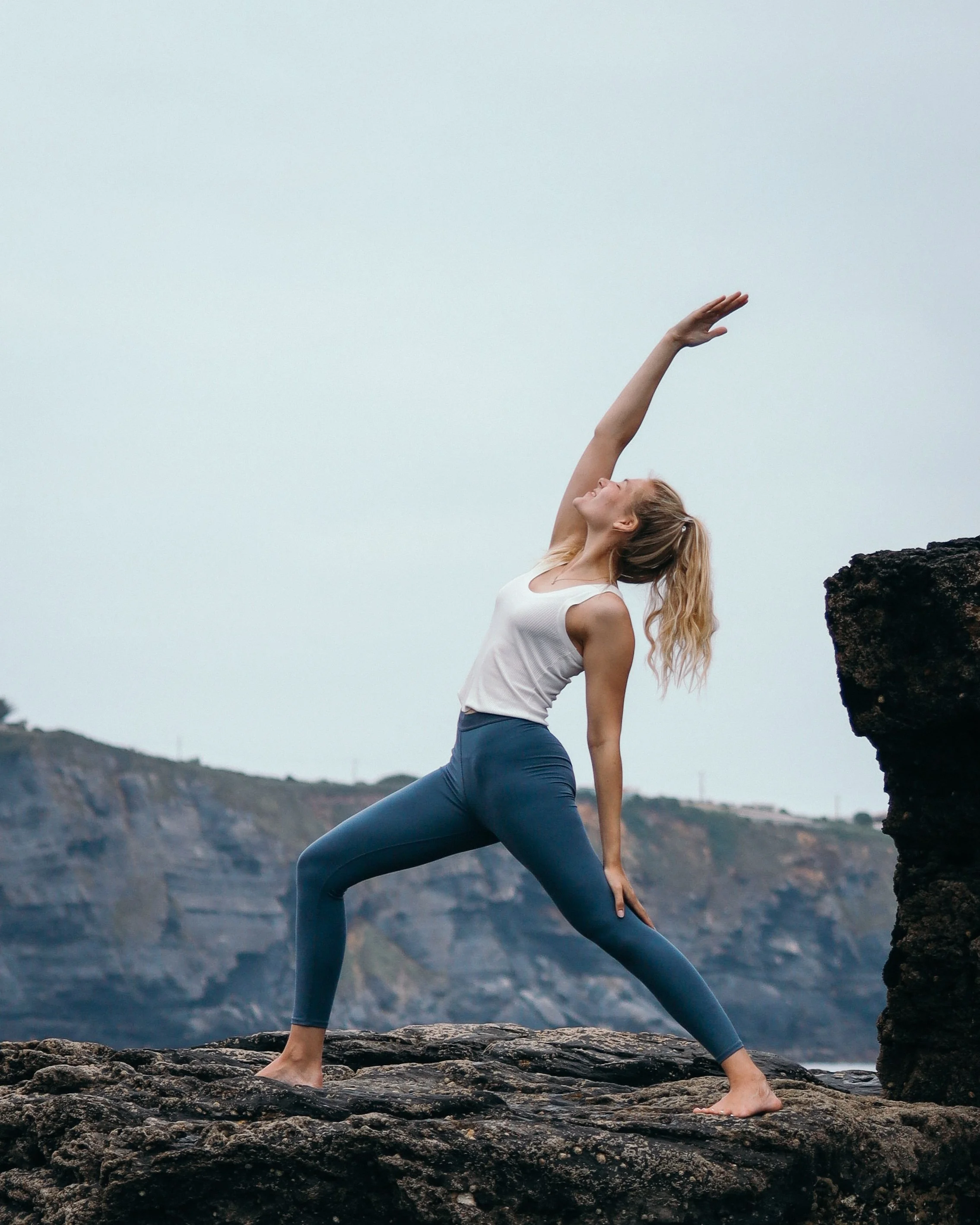Frau bei Yogaübung auf einer Felsenlandschaft am Meer, mit Hüftöffnung, Blick nach oben, bewölkter Himmel in der Ferne.