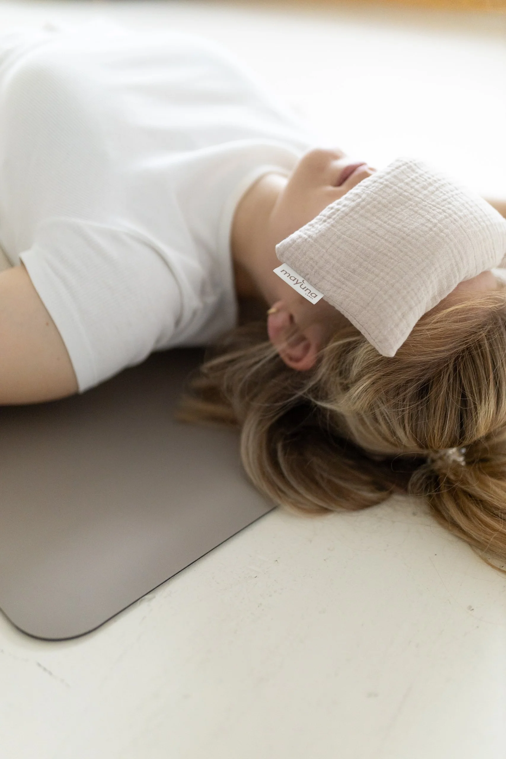 Frau liegt entspannt mit weißen Schlafmasken auf den Augen auf einem Tisch, trägt weißes T-Shirt, braune Haar und Ohrstecker, die Schlafmaske trägt das Markenetikett 'mayuna'.