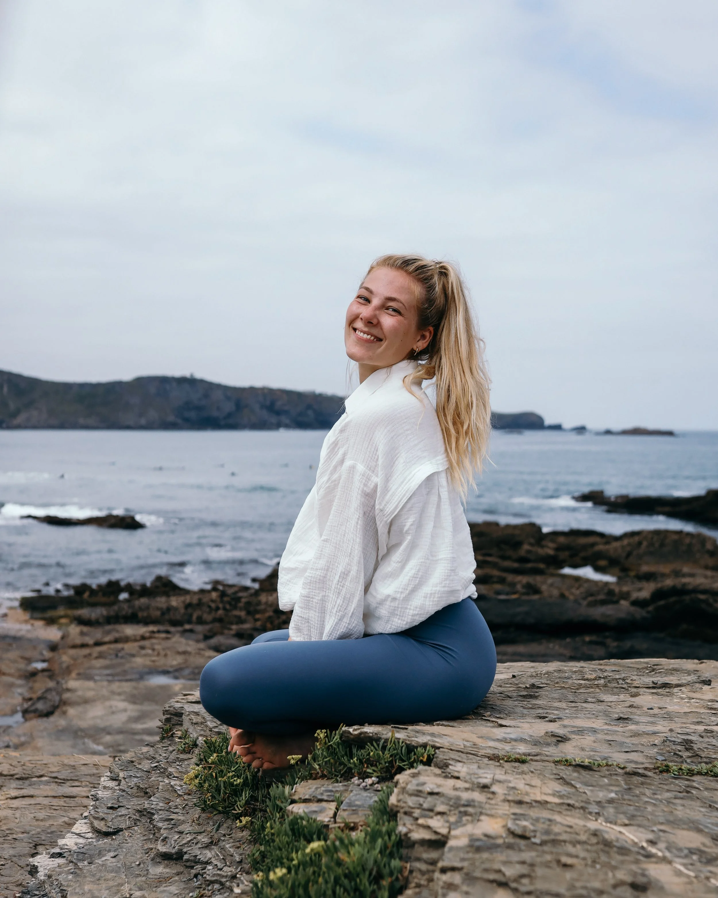 Eine lächelnde junge Frau sitzt im Schneidersitz auf einem Felsen am Meer, bekleidet mit weißem Hemd und blauen Hosen, im Hintergrund ist das Meer und ein bewölkter Himmel zu sehen.