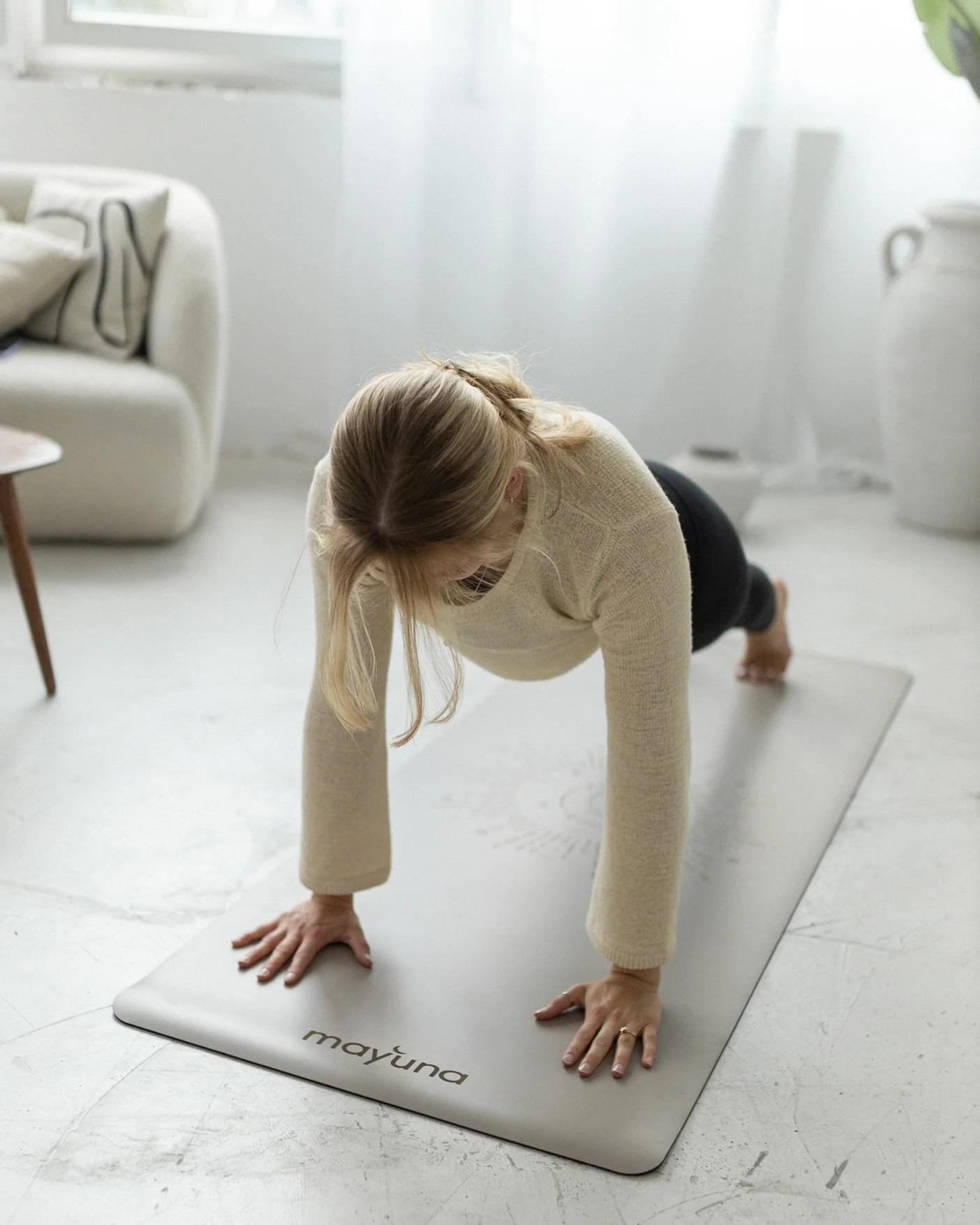 Frau macht Yoga-Übung im Wohnzimmer auf Yogamatte mit der Aufschrift 'mayüna'.