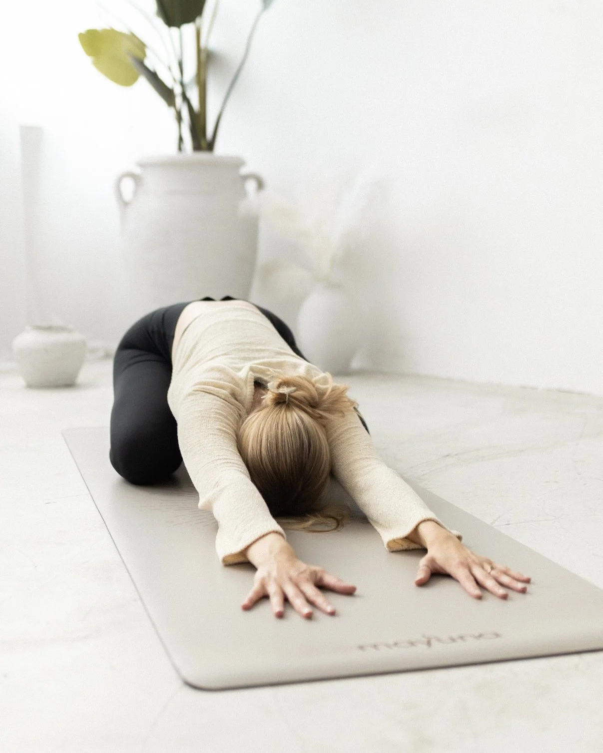Eine Frau macht einen Yogapose auf einer Yogamatte in einem hellen Raum mit weißen Wänden und großen Pflanzen.