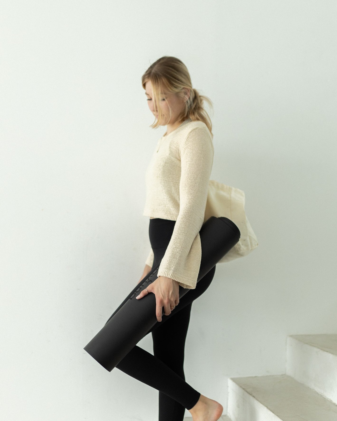 Frau in beige Pullover und schwarze Leggings macht Yoga auf weißen Stufen vor weißer Wand.