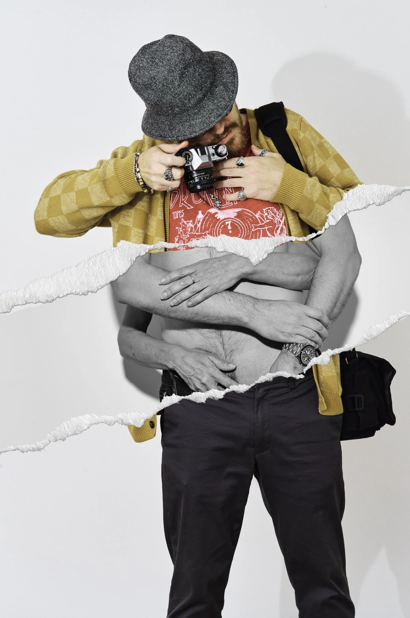A man in a mustard checkered jacket, red graphic t-shirt, and black pants is partially revealed by torn paper effects. His face is tilted down as he holds a camera near his face. He wears rings, bracelets, a watch, and a gray hat, with a black should