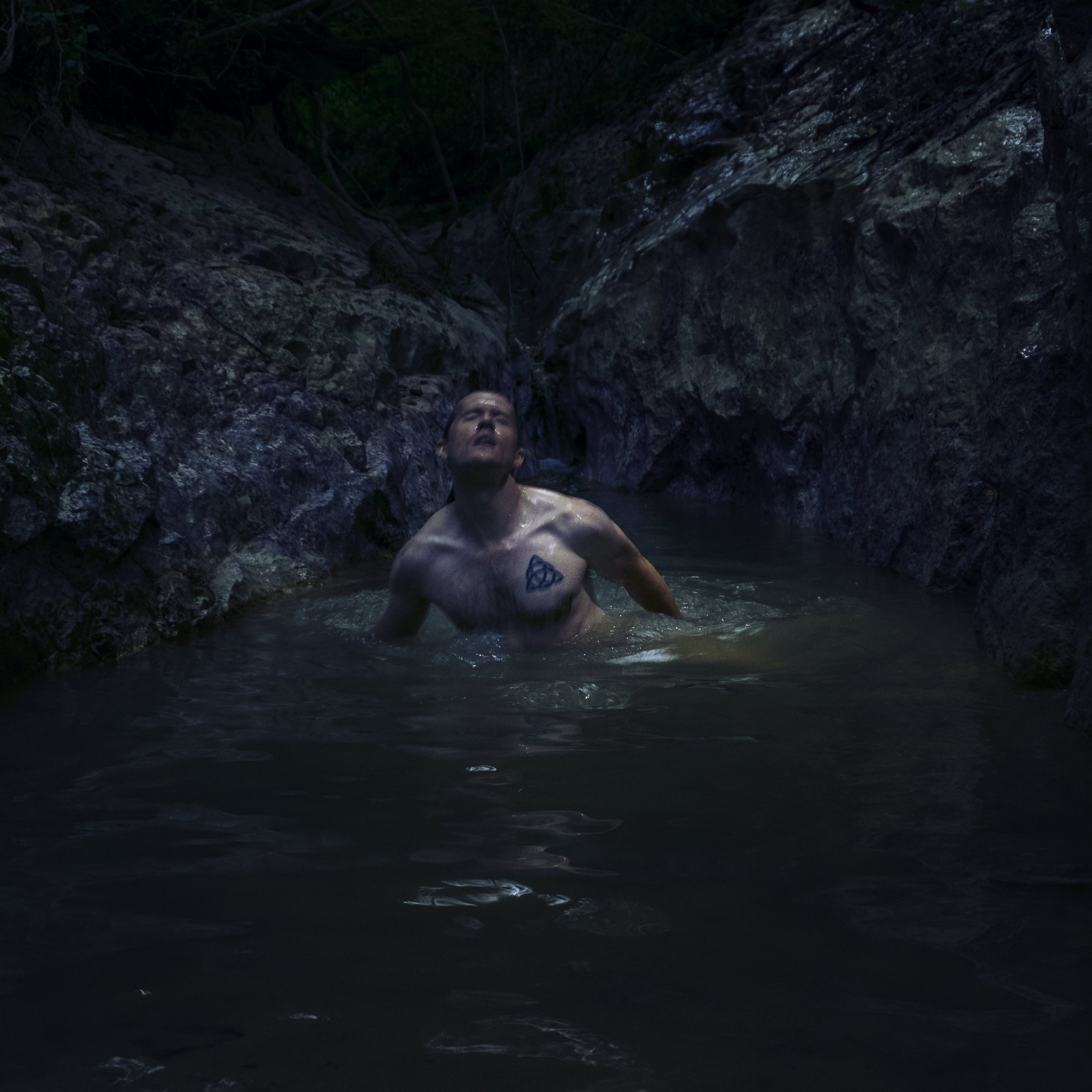 A person with a tattoo on his chest standing in a natural rocky water setting, with dark rocks and water surrounding him, and a waterfall in the background.
