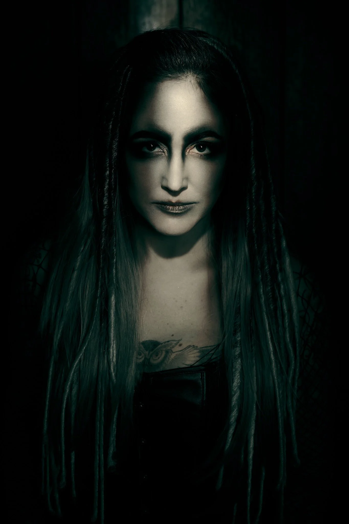 A woman with long hair, tattoos on her chest, and dark makeup, staring directly at the camera with a serious expression, dark background.