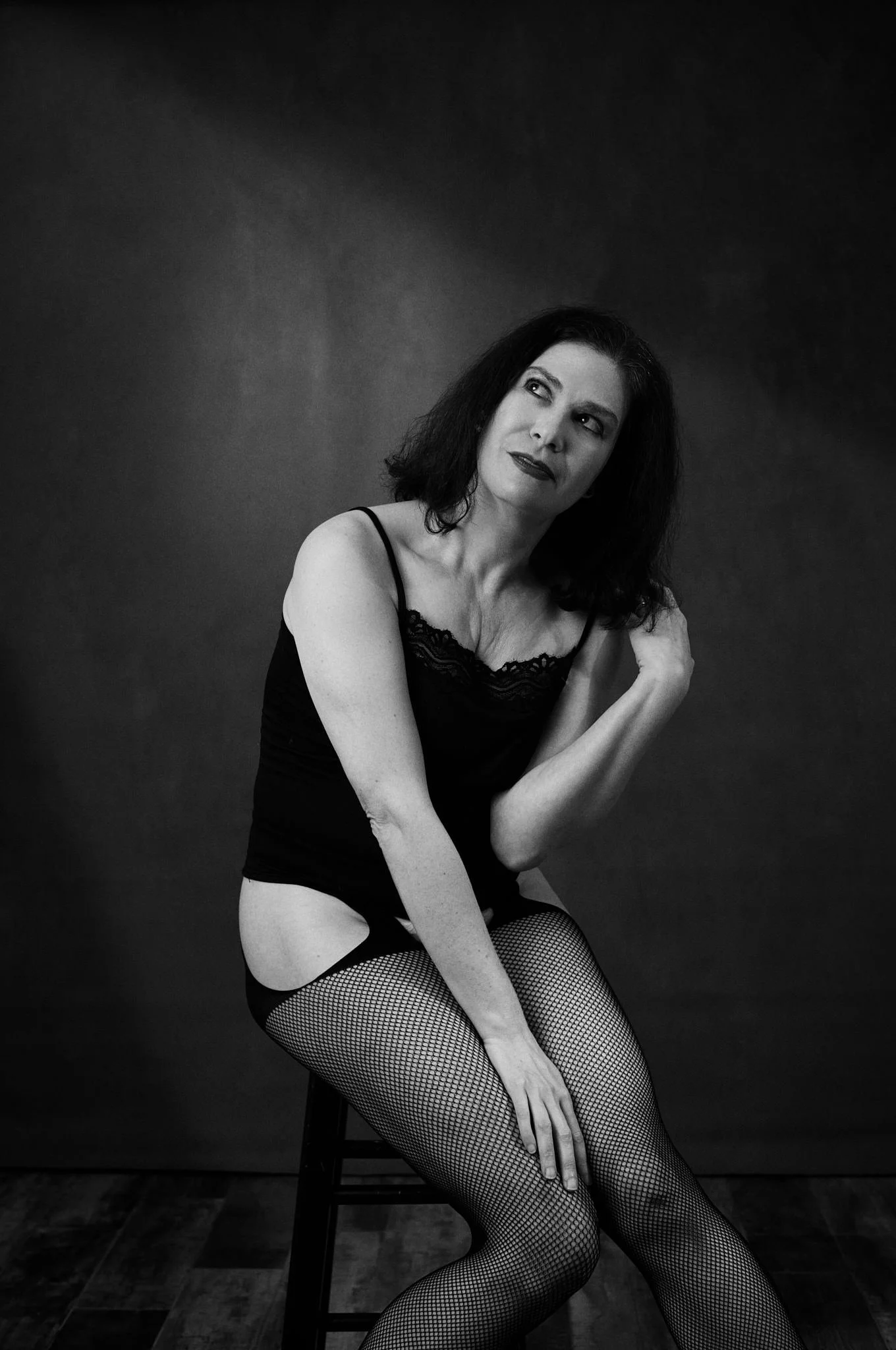 A black and white image of a woman wearing fishnets and a cammy. She's relaxed with a slight smile as she looks off camera and plays with her hair.